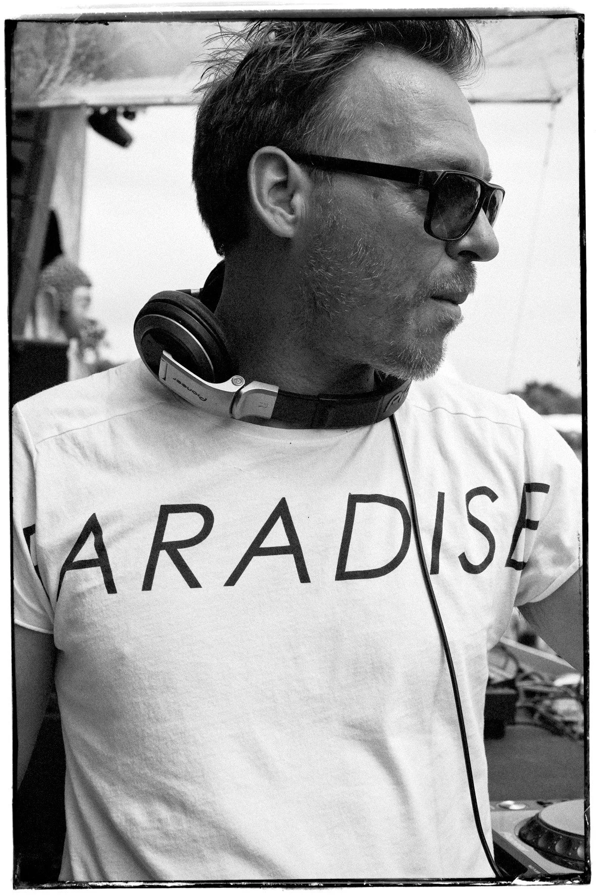 A electronic music artist with sunglasses, headphones around his neck, and a beard, wearing a T-shirt that says 'PARADISE' in large letters, standing outdoors.