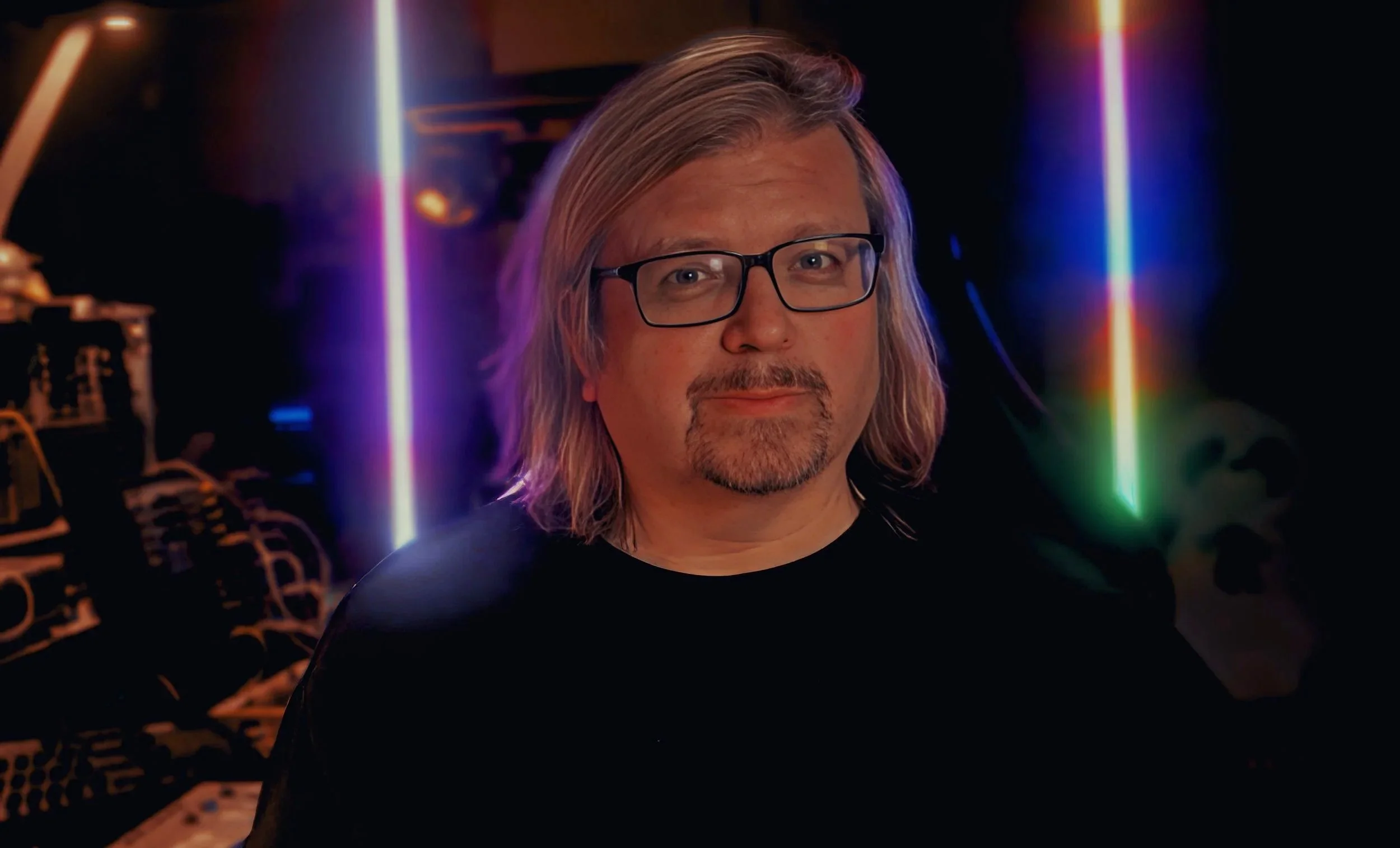 A music artist with shoulder-length gray hair, glasses, and a goatee, smiling in a dimly lit room with colourful vertical light streaks in the background.