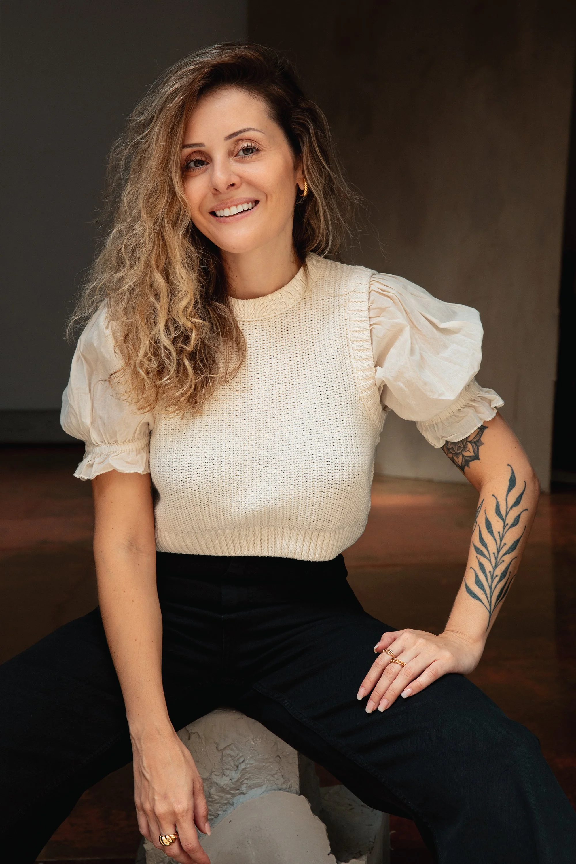 A woman with long, wavy blonde hair and tattoos on her right arm, wearing a cream-colored puff-sleeve top and black pants, seated on a stone sculpture in an indoor setting with a dark, neutral background.