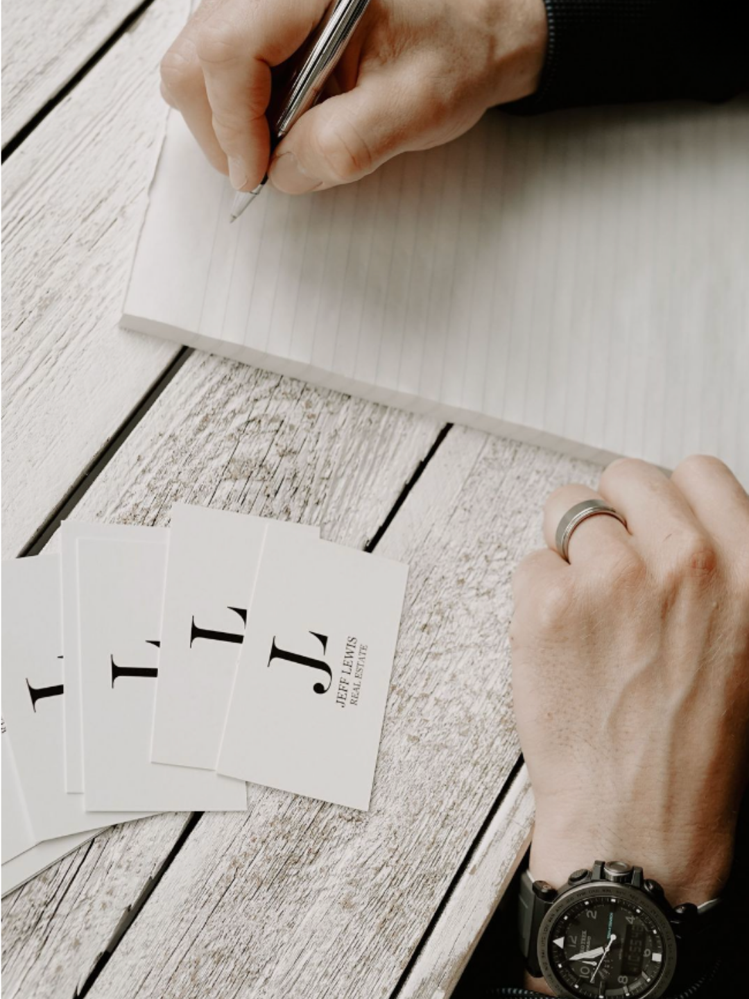 Person writing on lined paper with playing cards, including Jack of Clubs, on a rustic wooden table.
