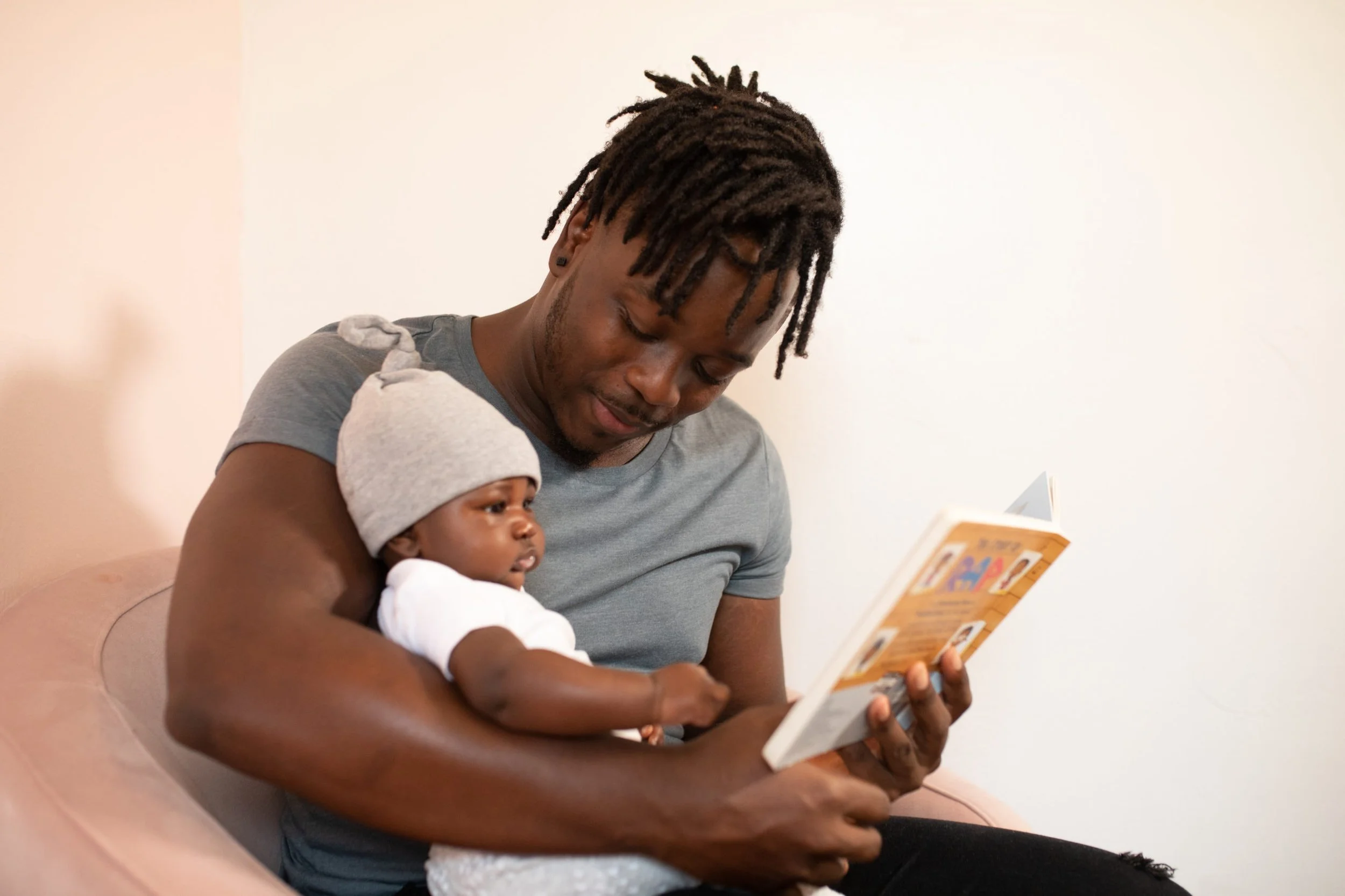 A father reads a book to his baby, who is wearing a gray hat and pointing at the pages.
