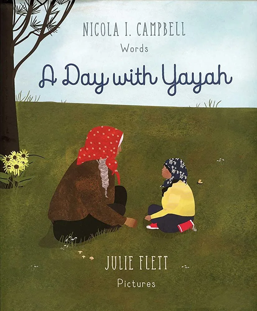A First Nations grandmother and granddaughter sit in the grass picking mushrooms and talking to one another.