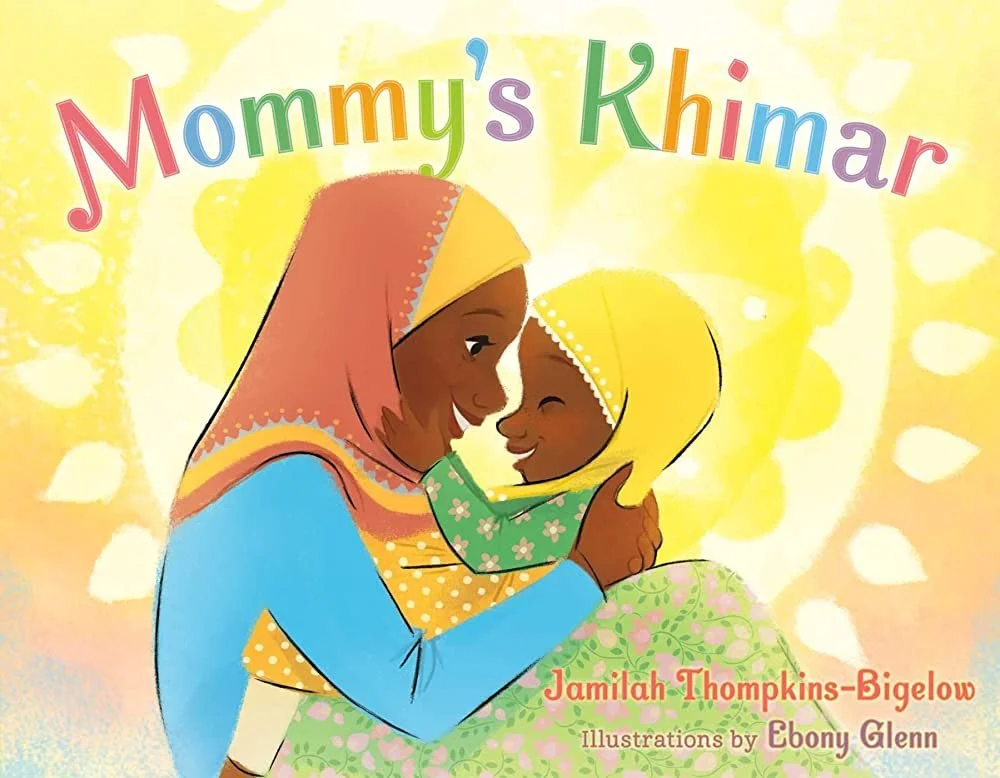 A young Muslim girl sits in her mother’s lap, wrapped in her mother's Khimar as they smile at one another.