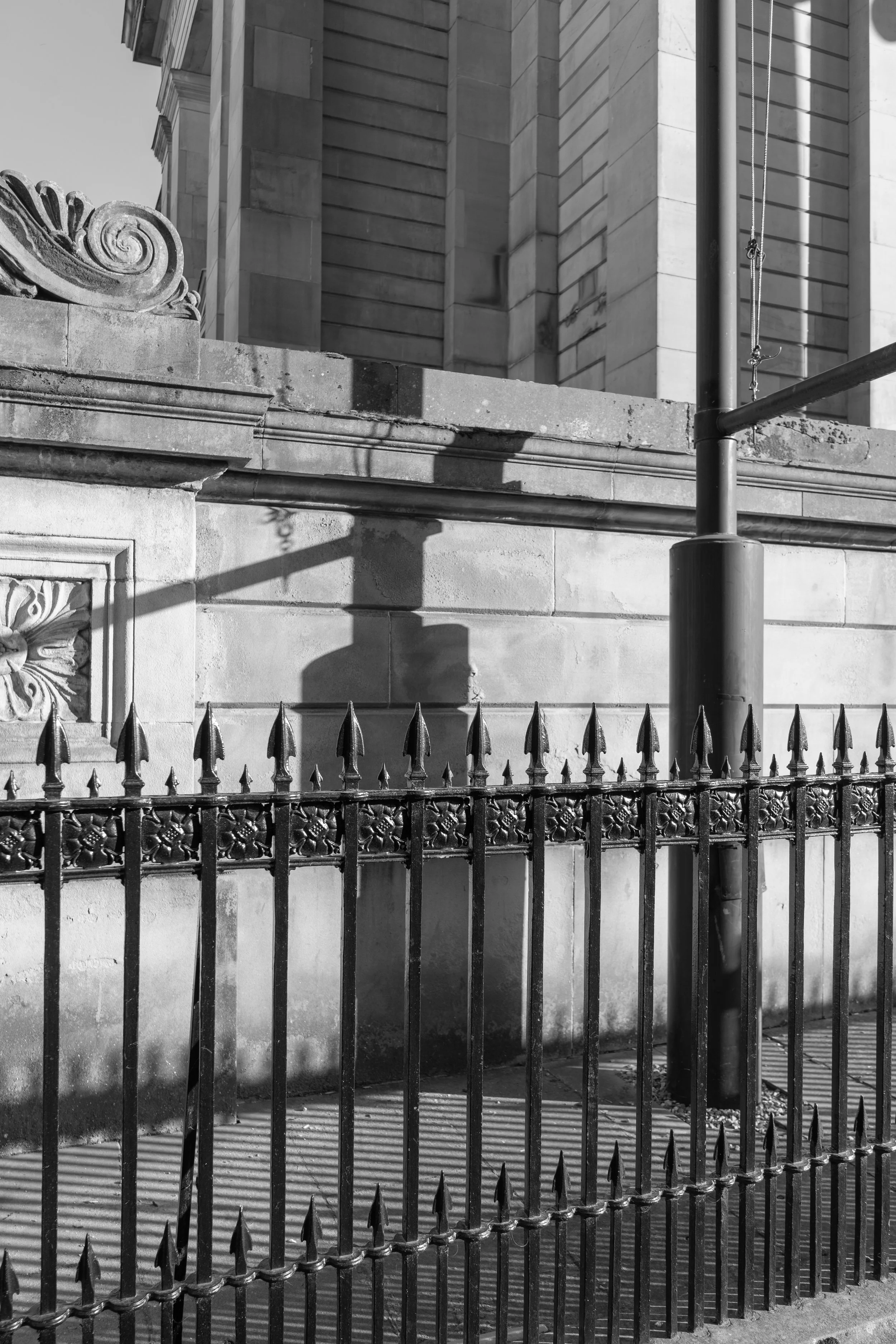 Black and white photo of a metal fence with pointed spikes, set in front of a stone wall with decorative architectural elements, and a building with a stone facade in the background. situated in Edinburgh