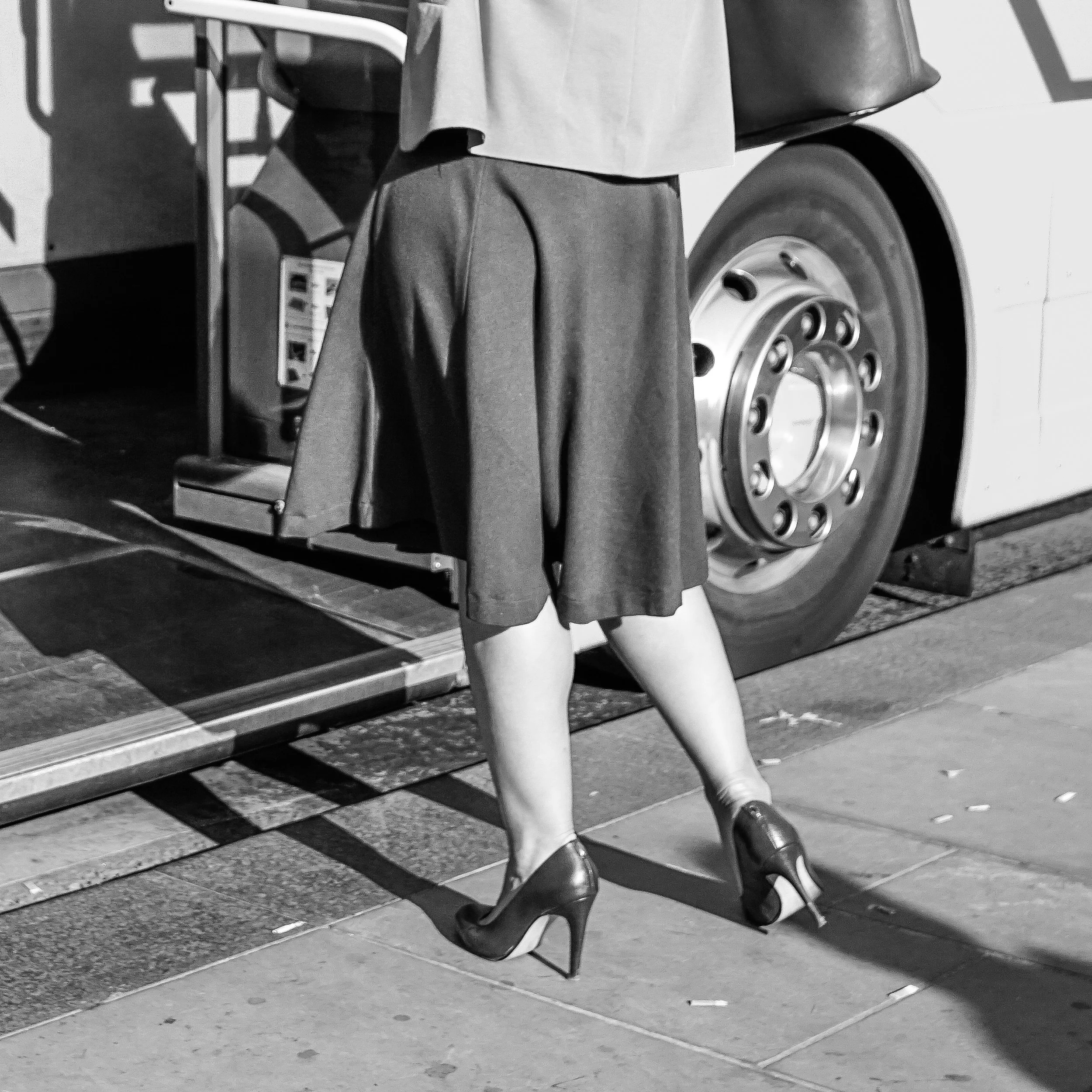 A person with a coat and shoes standing next to a large vehicle, possibly a bus or truck, with its wheel and part of the body visible, on Waverley Bridge, Edinburgh.