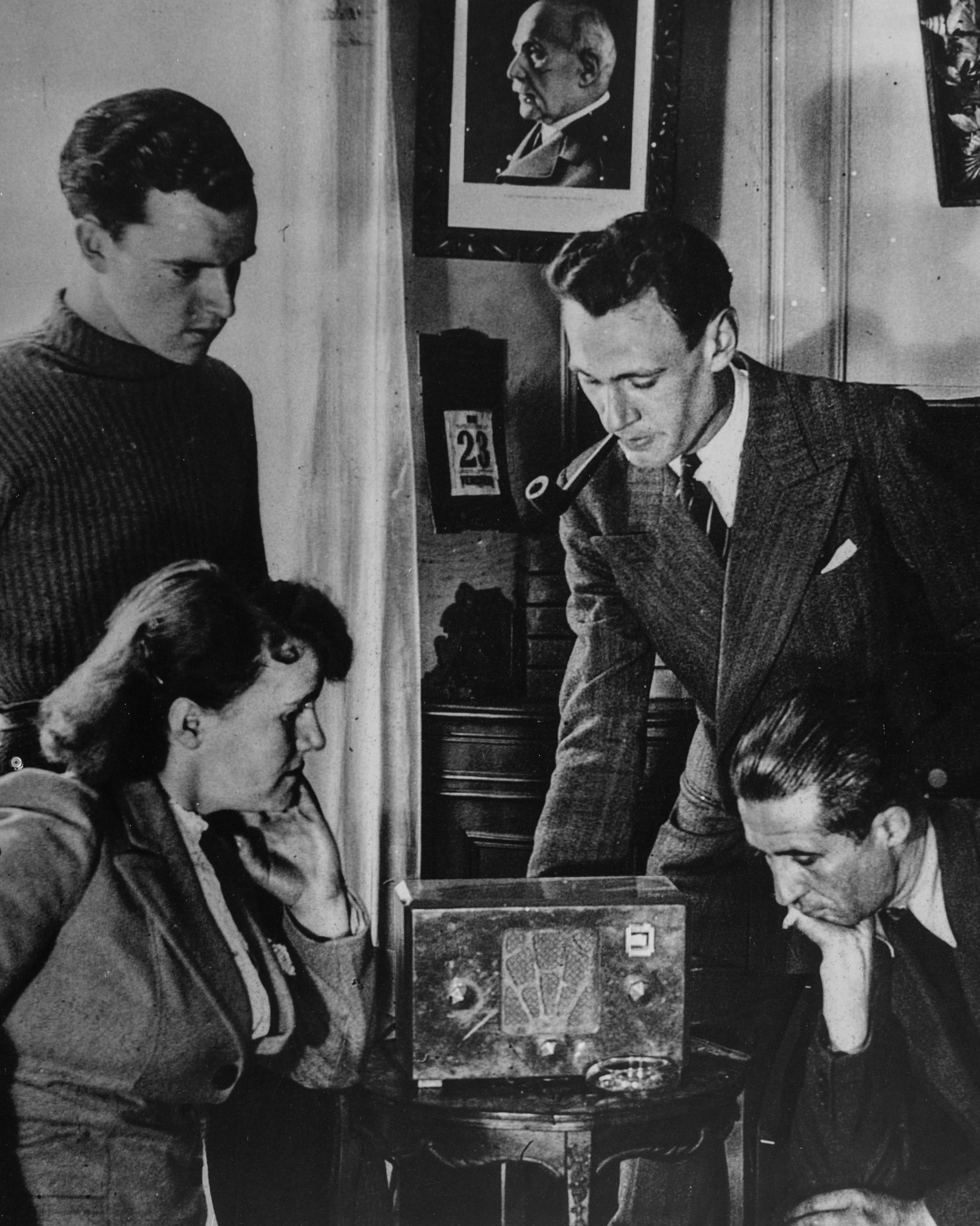 Black and white photo of four young individuals playing chess at a table. There are framed pictures on the wall, including a profile portrait of a man in a military uniform. The scene appears to be indoors.