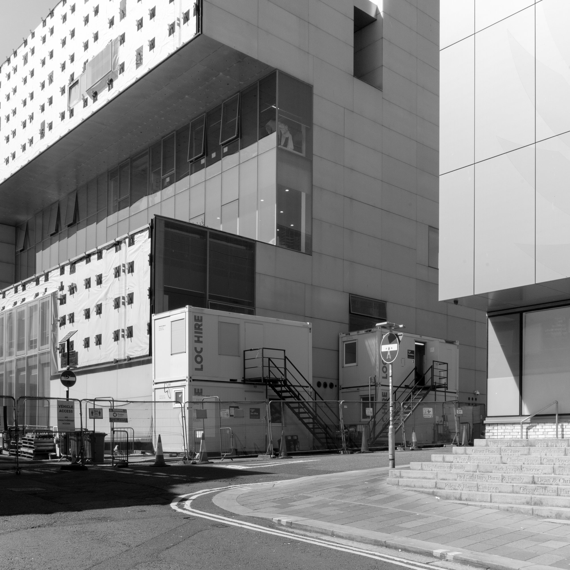 Construction site with portable offices and safety barriers outside a modern building with large glass windows.