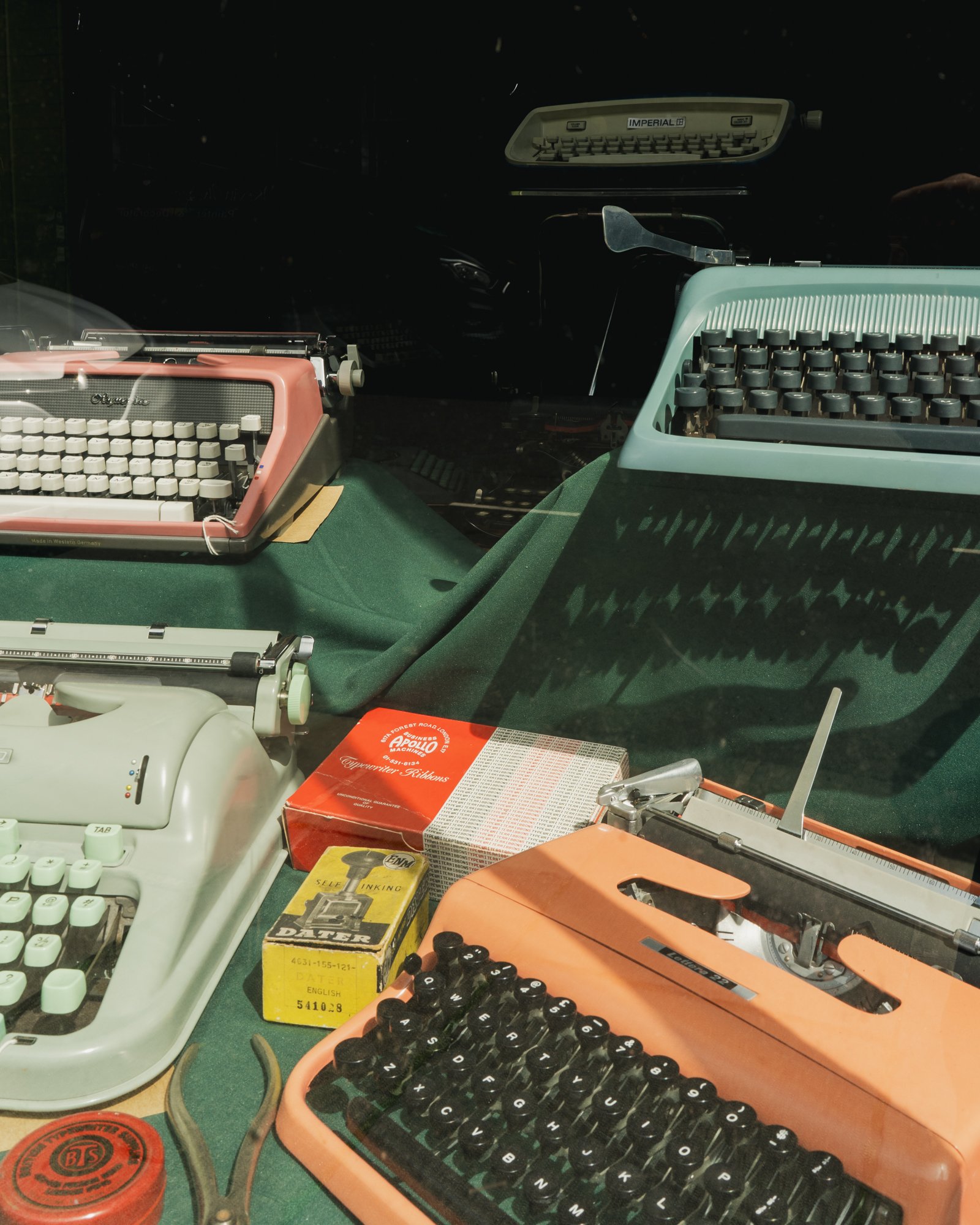 A display of vintage typewriters, including a pink one in the foreground and a green one in the background, on a green tablecloth with various old office supplies around.