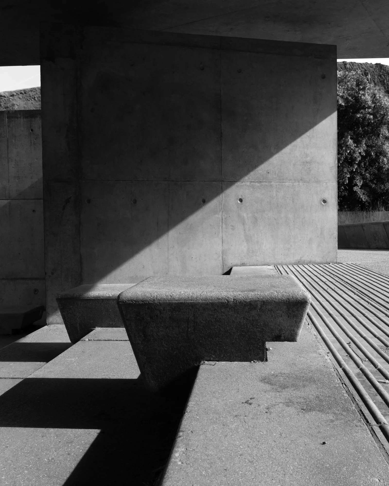 Black and white photograph of the Scottish Parliament Building with geometric shapes and shadows. A large concrete wall with circular holes, part of an overhanging roof, and a large concrete bench in the foreground.