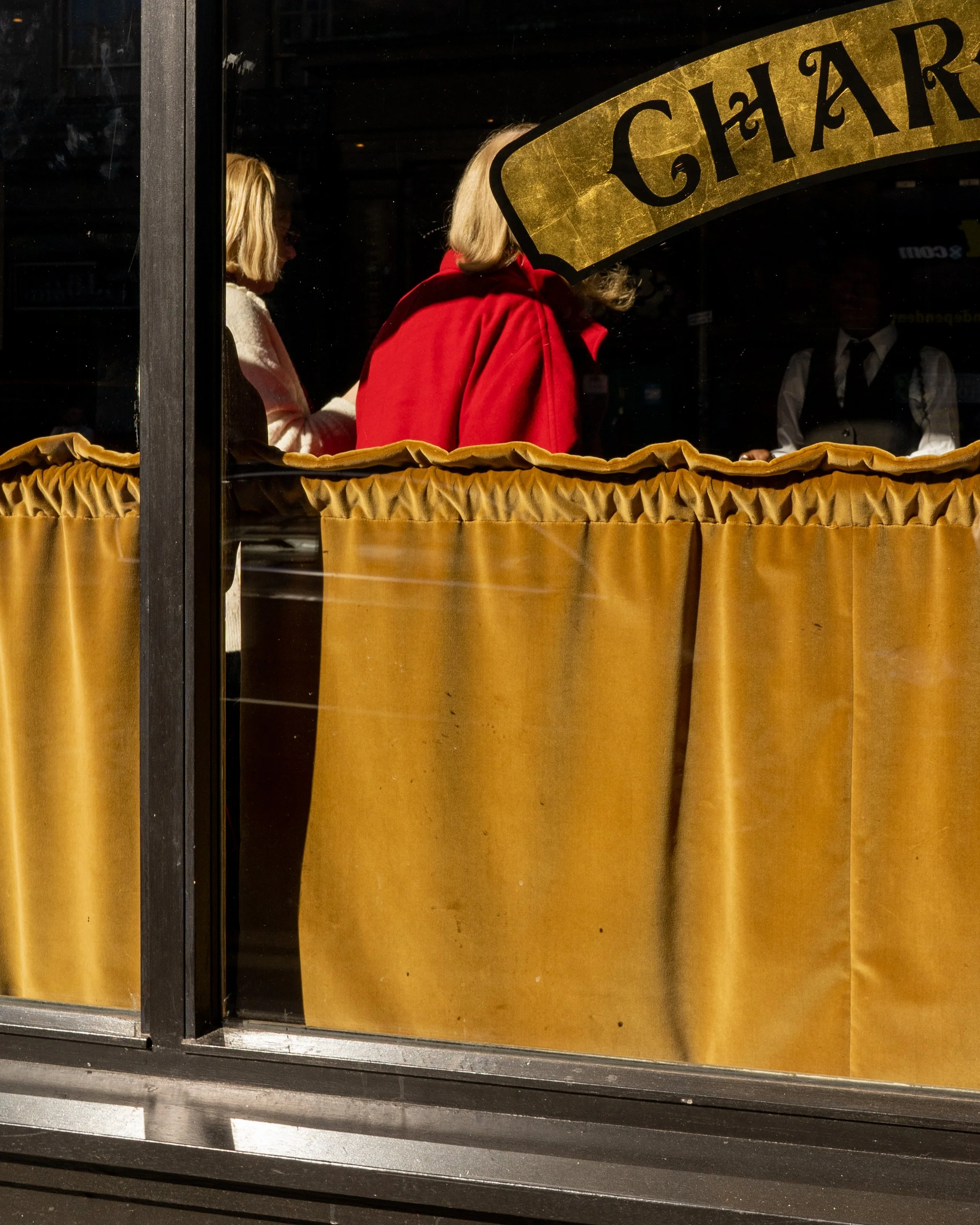 People visible through a restaurant window with a gold and black sign partially reading 'CH' and an orange curtain covering the bottom part of the window.