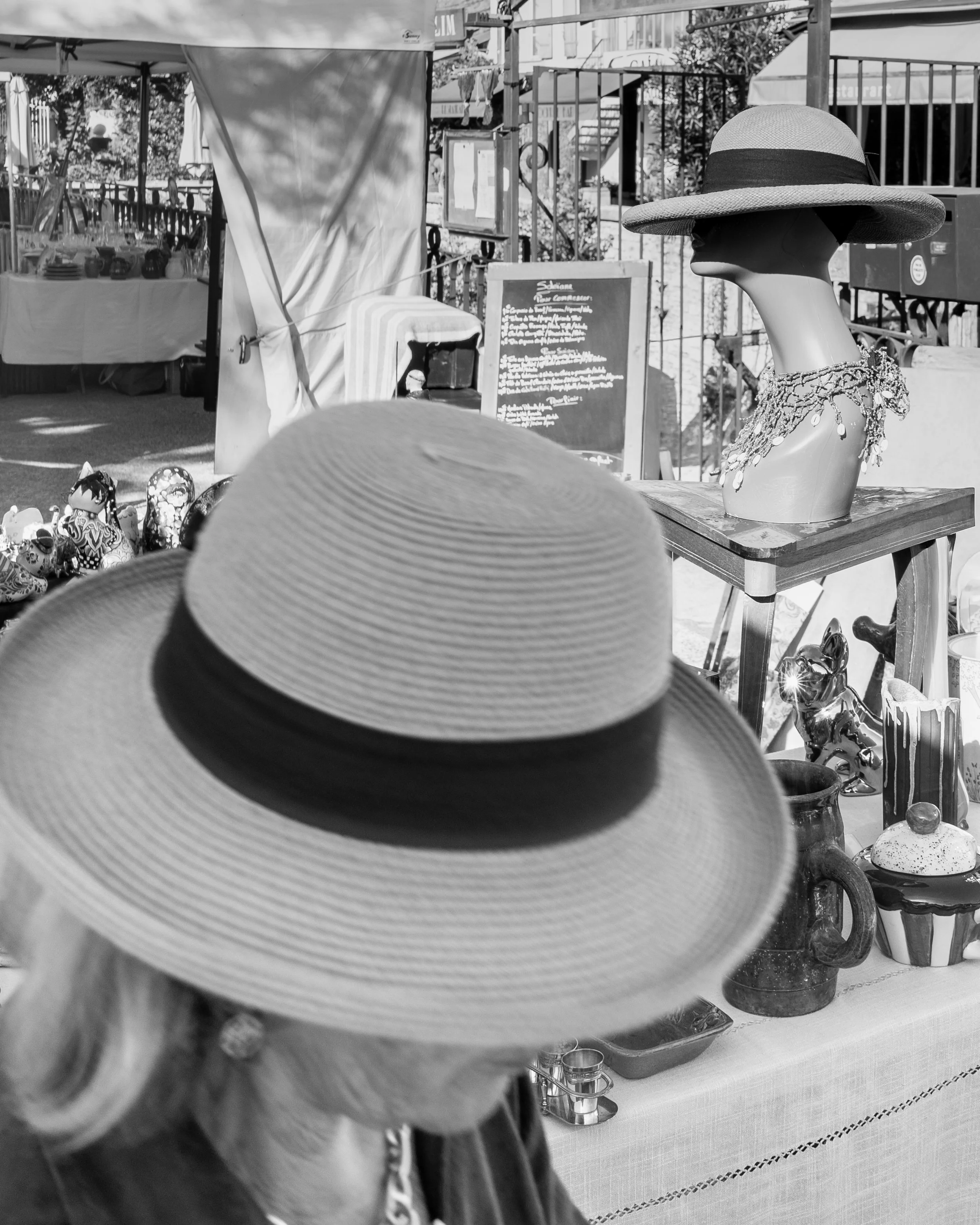 Black and white photo taken at an outdoor market or craft fair. In the foreground, a person wearing a wide-brimmed hat is holding a straw hat with a black band, partially obscuring the view. Behind, there is a display of various decorative items and knick-knacks, including small figurines and ceramic pieces. A mannequin bust with a wide-brimmed hat and beaded necklace is prominently placed on a table. Further in the background, there are tents, a chalkboard menu, and a fenced area, indicating a busy outdoor shopping or market scene.