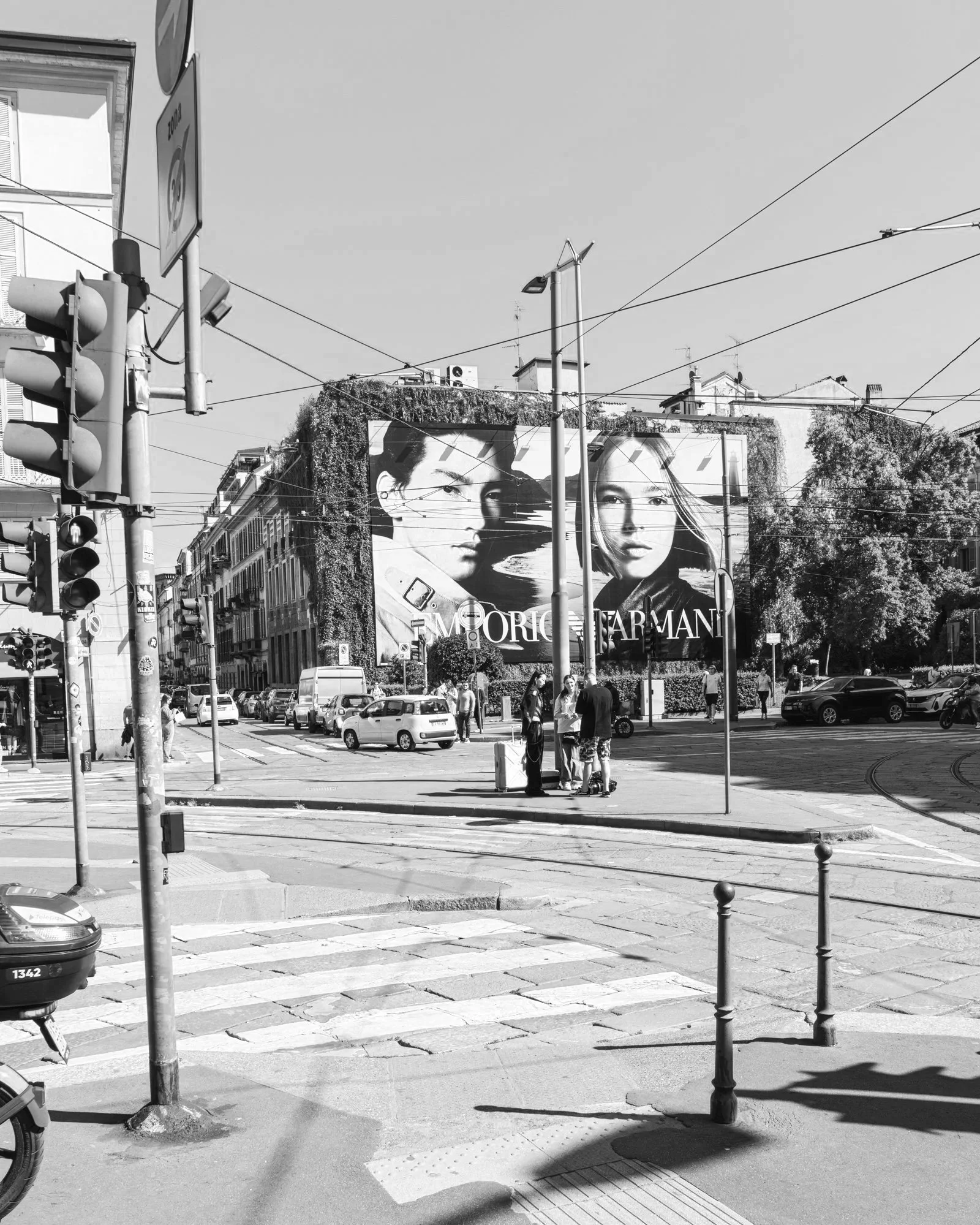Black and white street scene with tram tracks, traffic signals, cars, pedestrians, buildings, and a large billboard featuring fashion models.