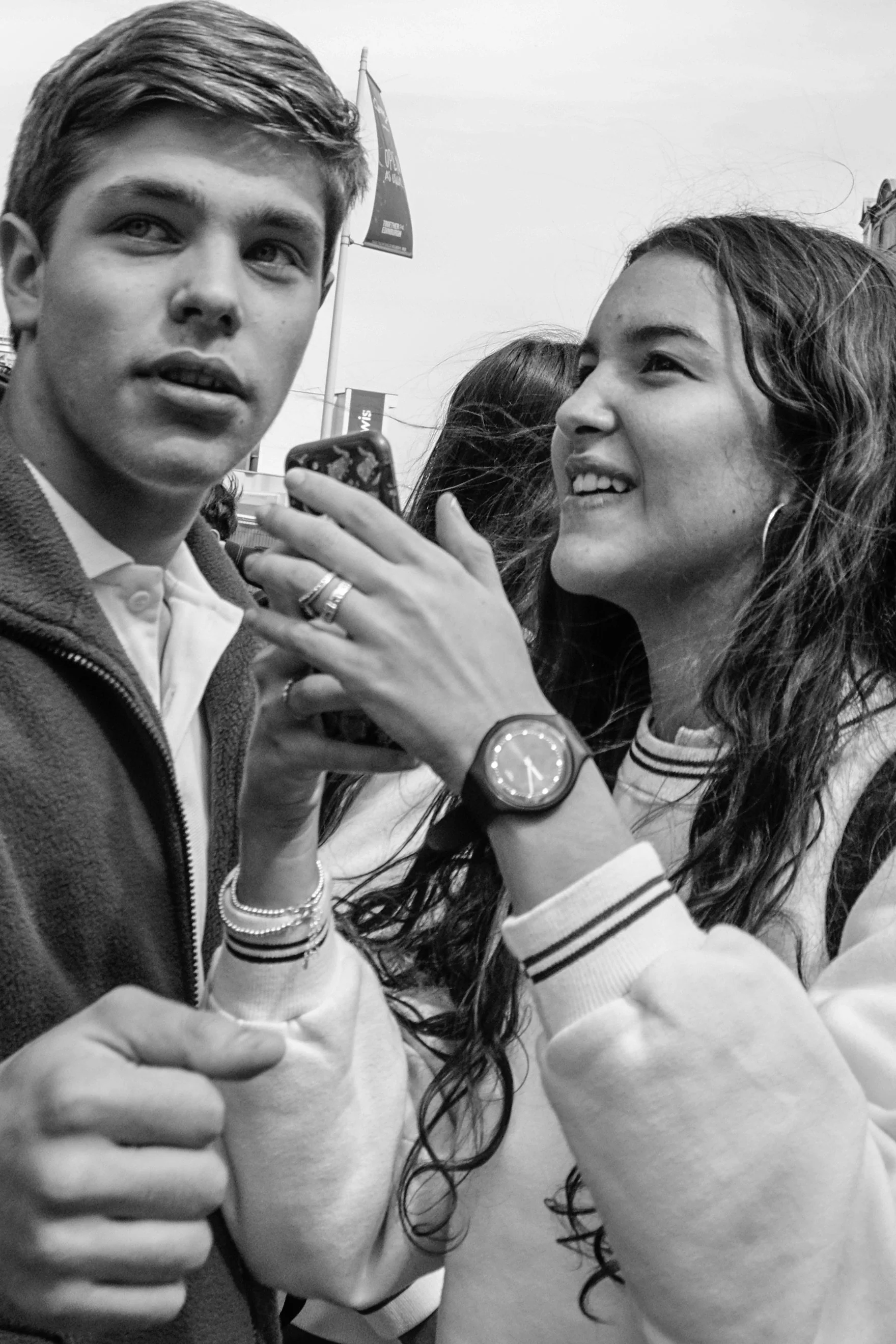 Two young people, a man and a woman, are taking a selfie together outdoors. The woman is smiling, and the man is holding a phone. They are dressed casually, with the woman wearing a striped sweatshirt and the man wearing a hoodie.