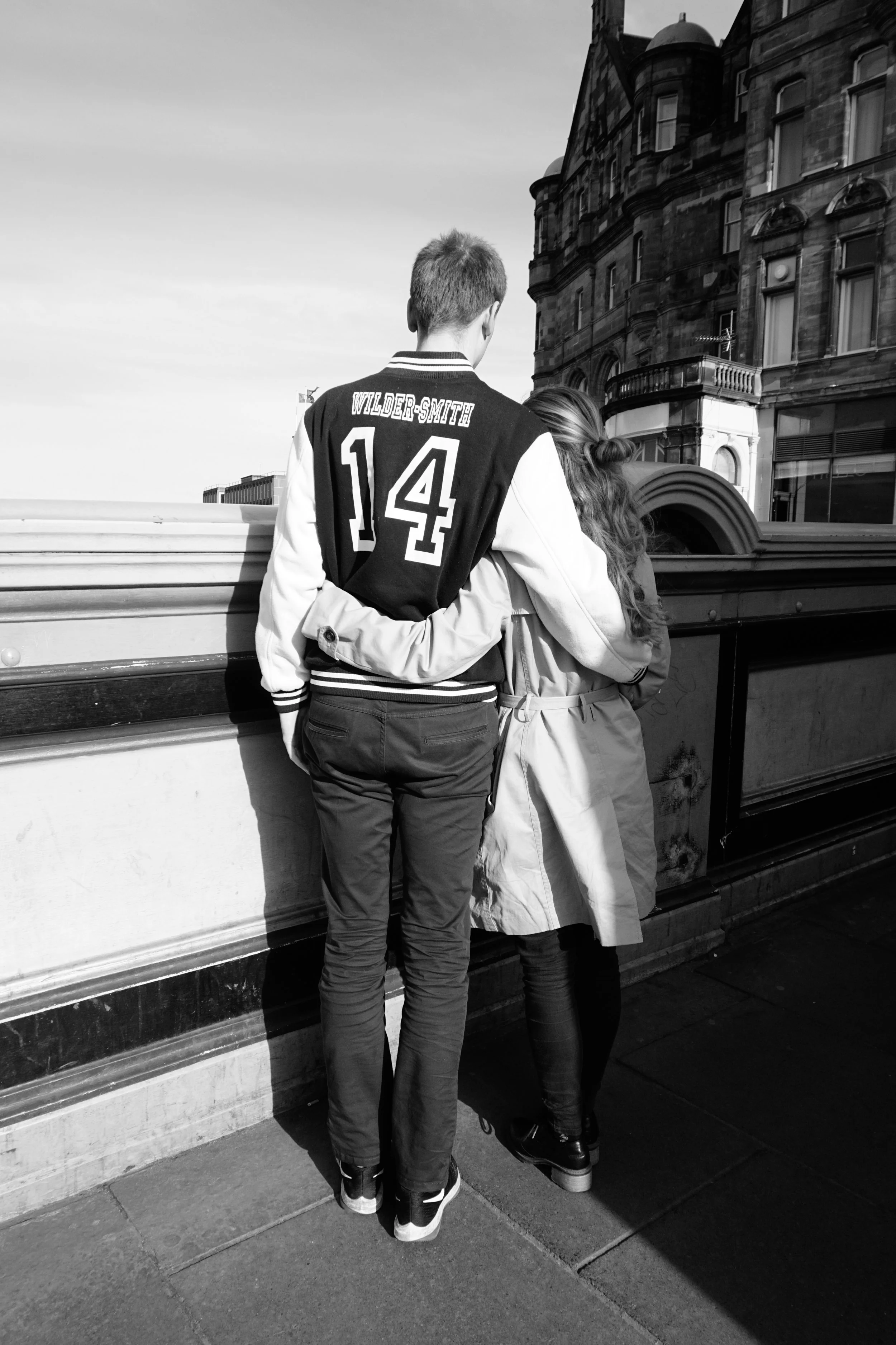 A young man and woman standing close together on a city street, seen from behind. The man is wearing a black varsity jacket with 'Wilder-Smith 14' on the back, and the woman has long curly hair and is wearing a coat. They are leaning against a bridge railing with a historic building in the background.