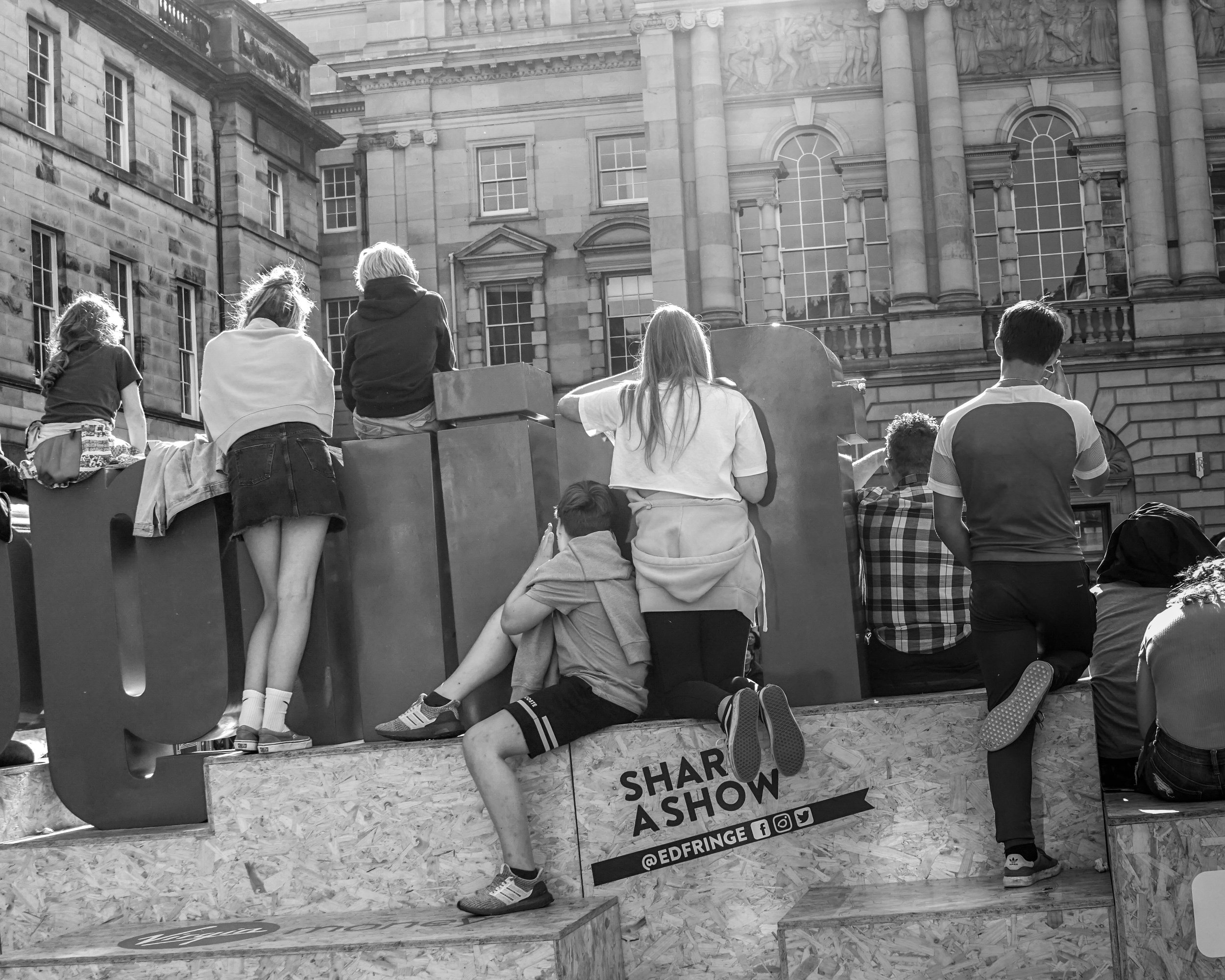 People gathered around a booth at the Edge Fringe Festival, with a historic building in the background. Some are sitting and others are standing, with a backdrop that has event information and social media icons.