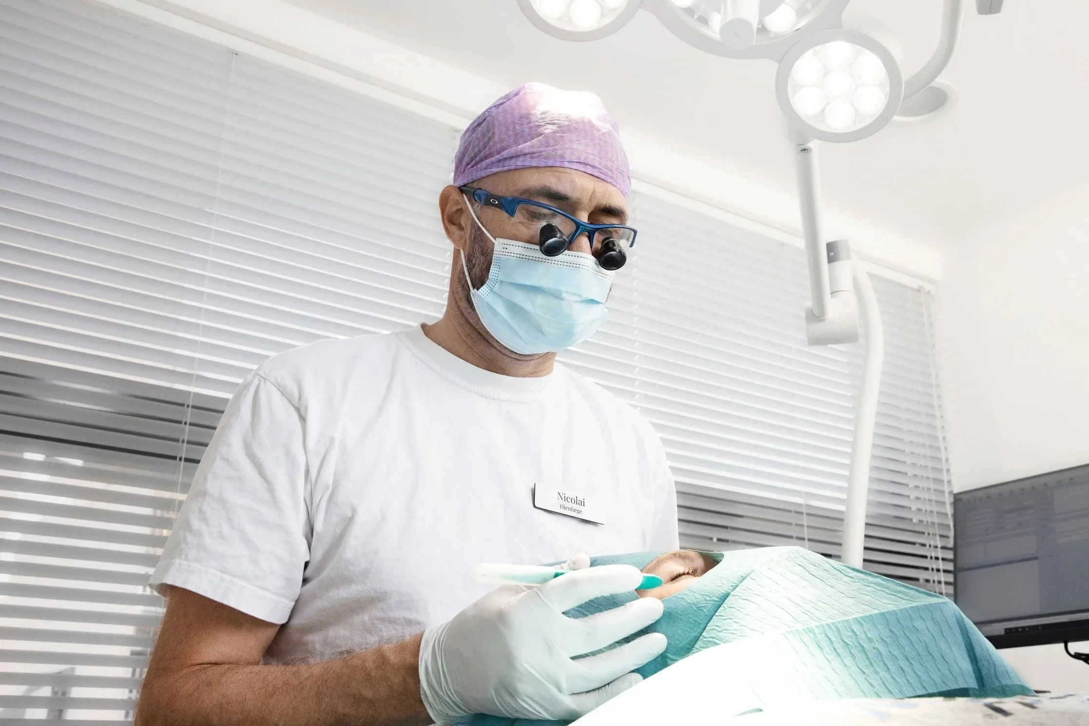 En læge iført scrub, maske og beskyttelsesbriller arbejder med en patient på operationsbordet. Der er medicinsk udstyr og en monitor i baggrunden.