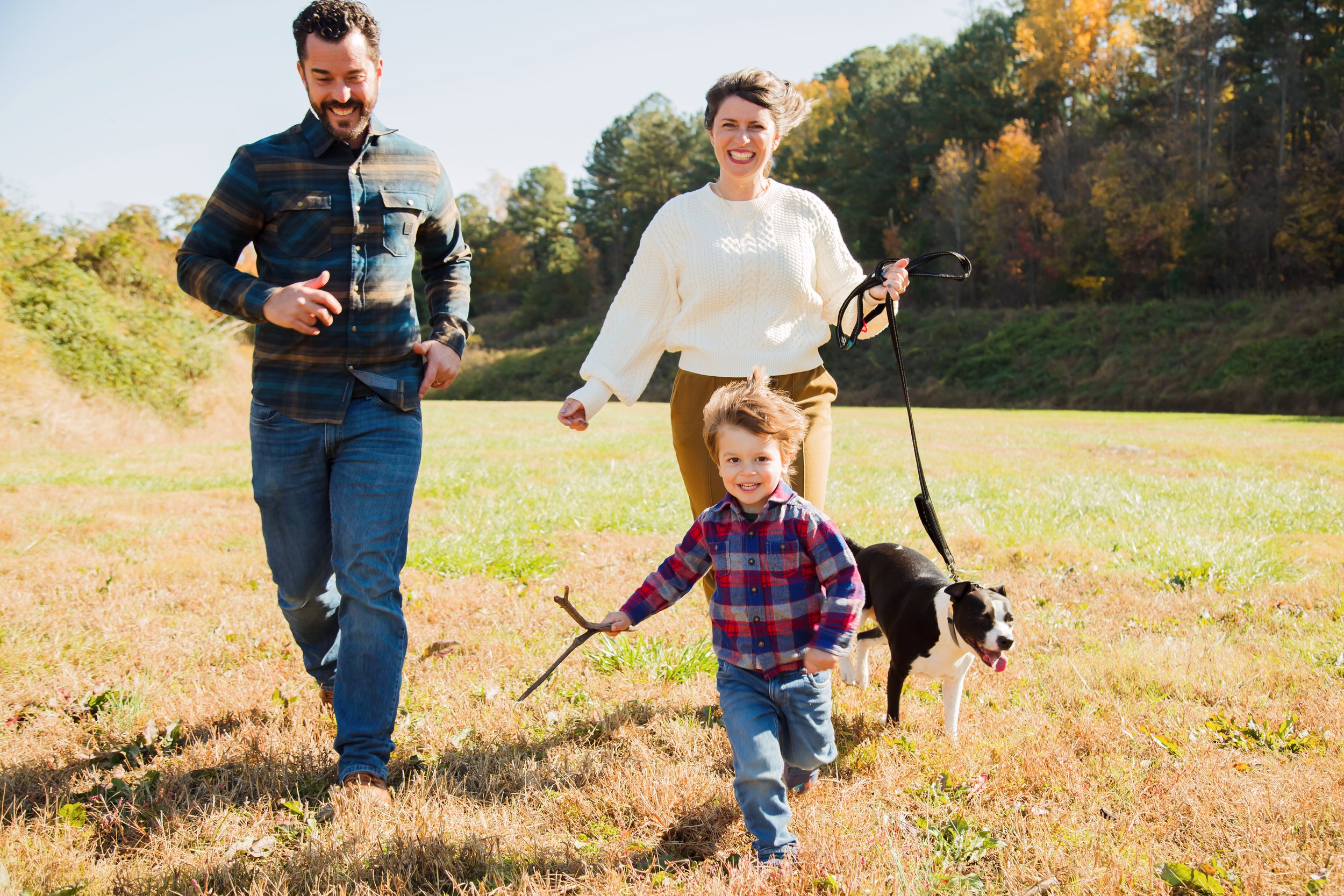 Family running towards the camera smiling - father, mother, and son. Mother is holding a leash with their black and white dog. Autumnal background.