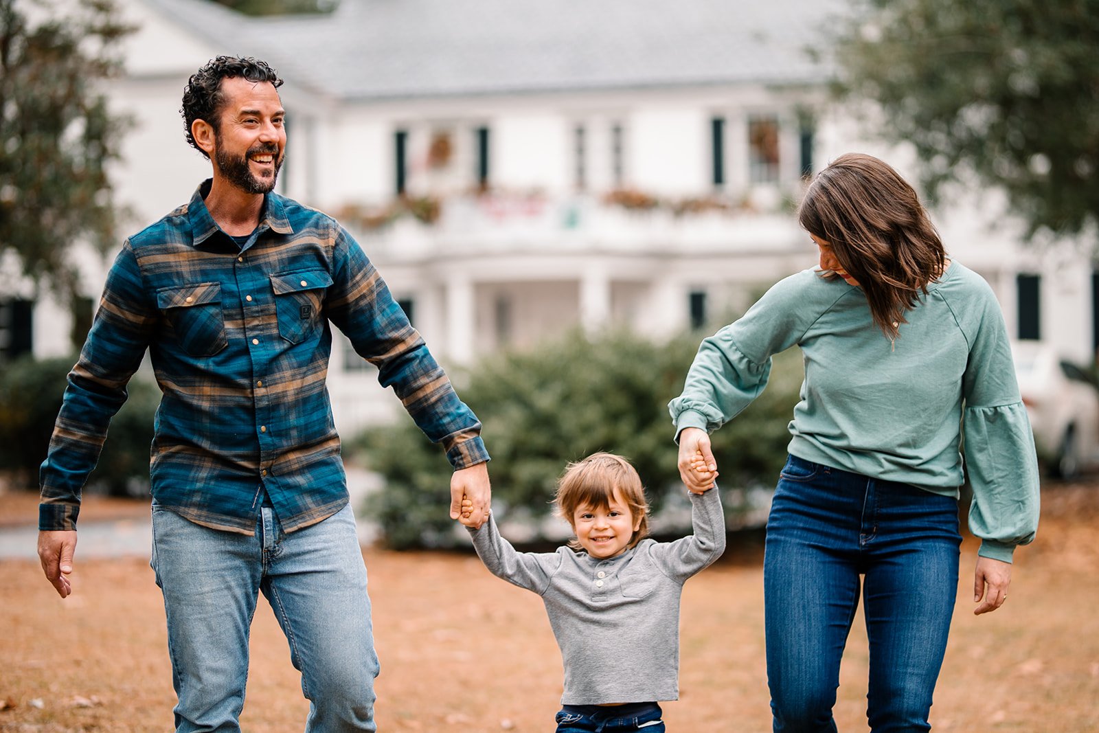 Father and mother holding son's hands, with the son in the middle as if he is about to be swung. White colonial style house blurred in the background.