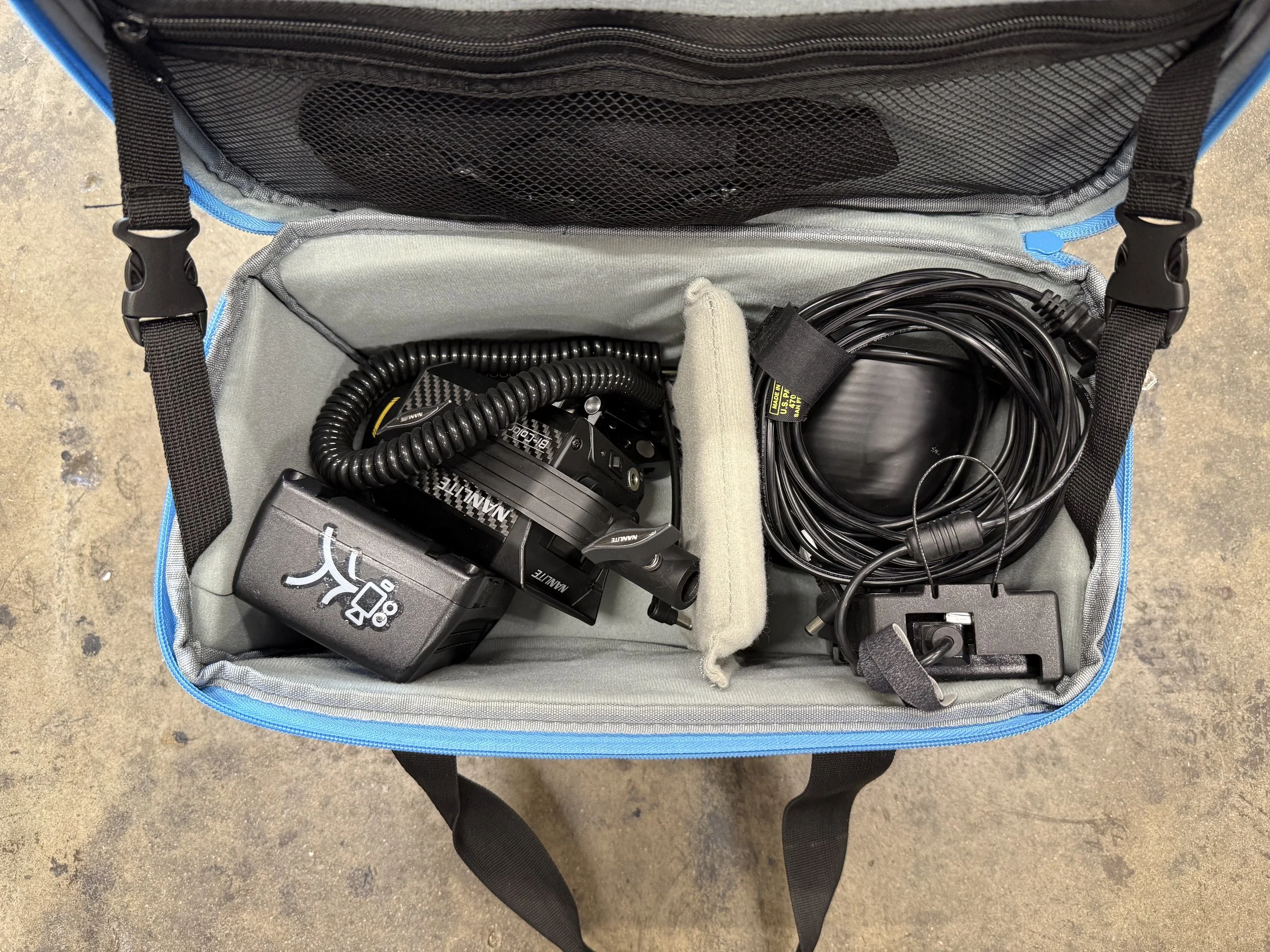 Open blue and gray carrying case with camera equipment, including a black Nikon camera, a battery charger, and coiled cables inside.