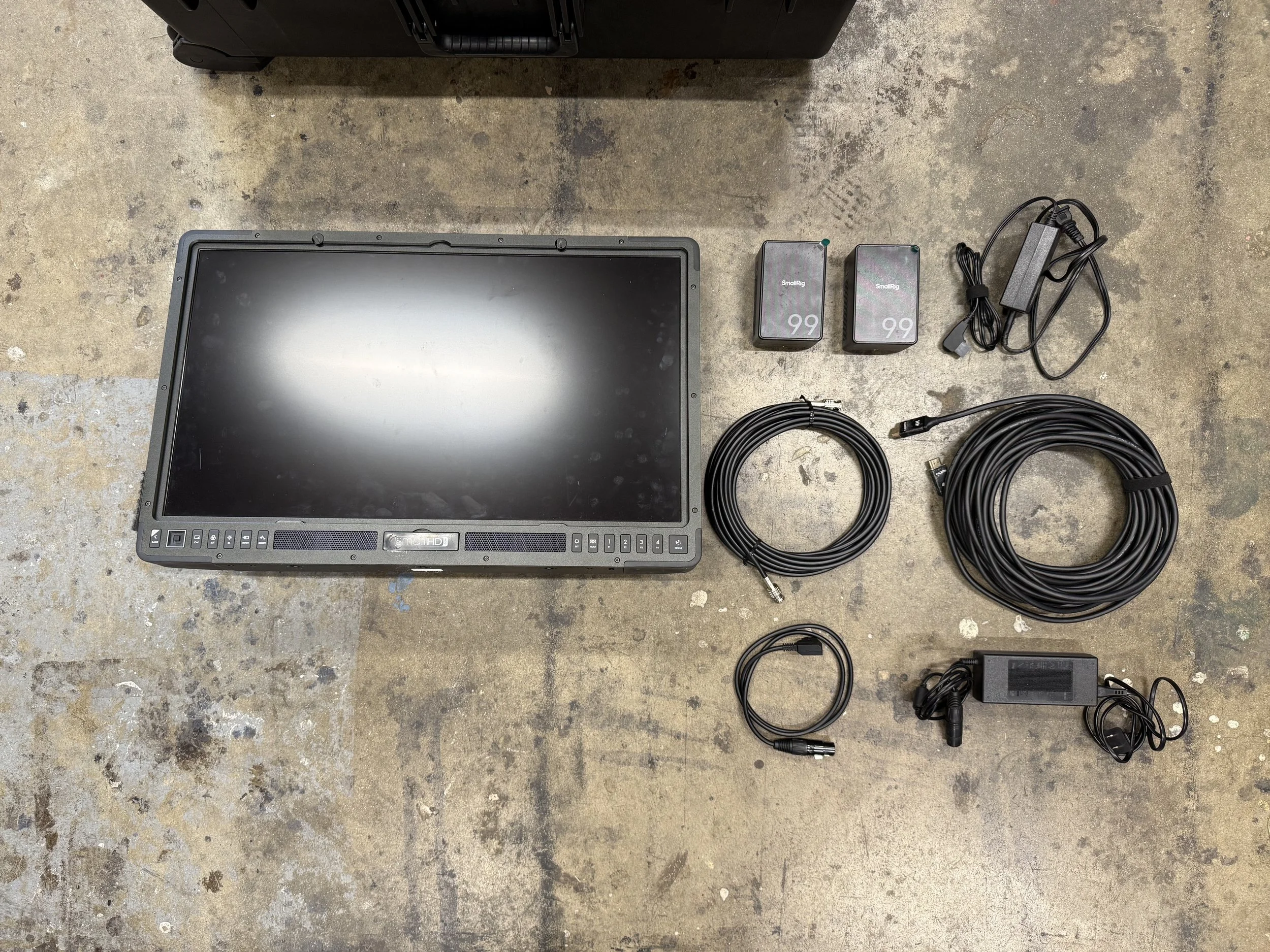 A large monitor with buttons below the screen, two wireless microphone packs labeled '99', a power adapter, two long black cables, a smaller black round cable, and a power brick with attached cables all arranged on a concrete floor.