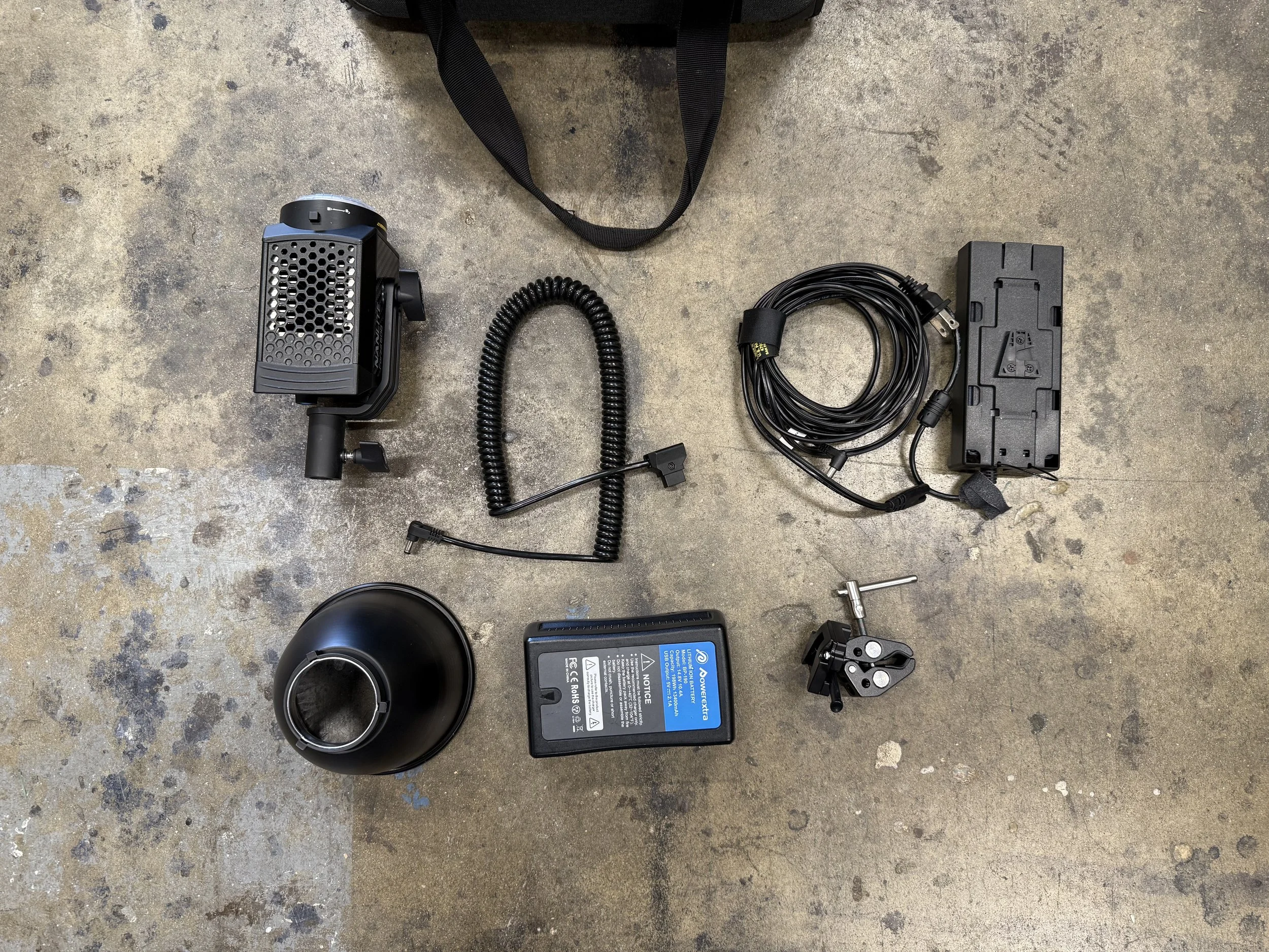 A collection of electronic and mechanical equipment laid out on a concrete floor, including a black and grey device, a coiled cable, a power supply, an adjustable mount, a black spherical object, and a black carrying bag.