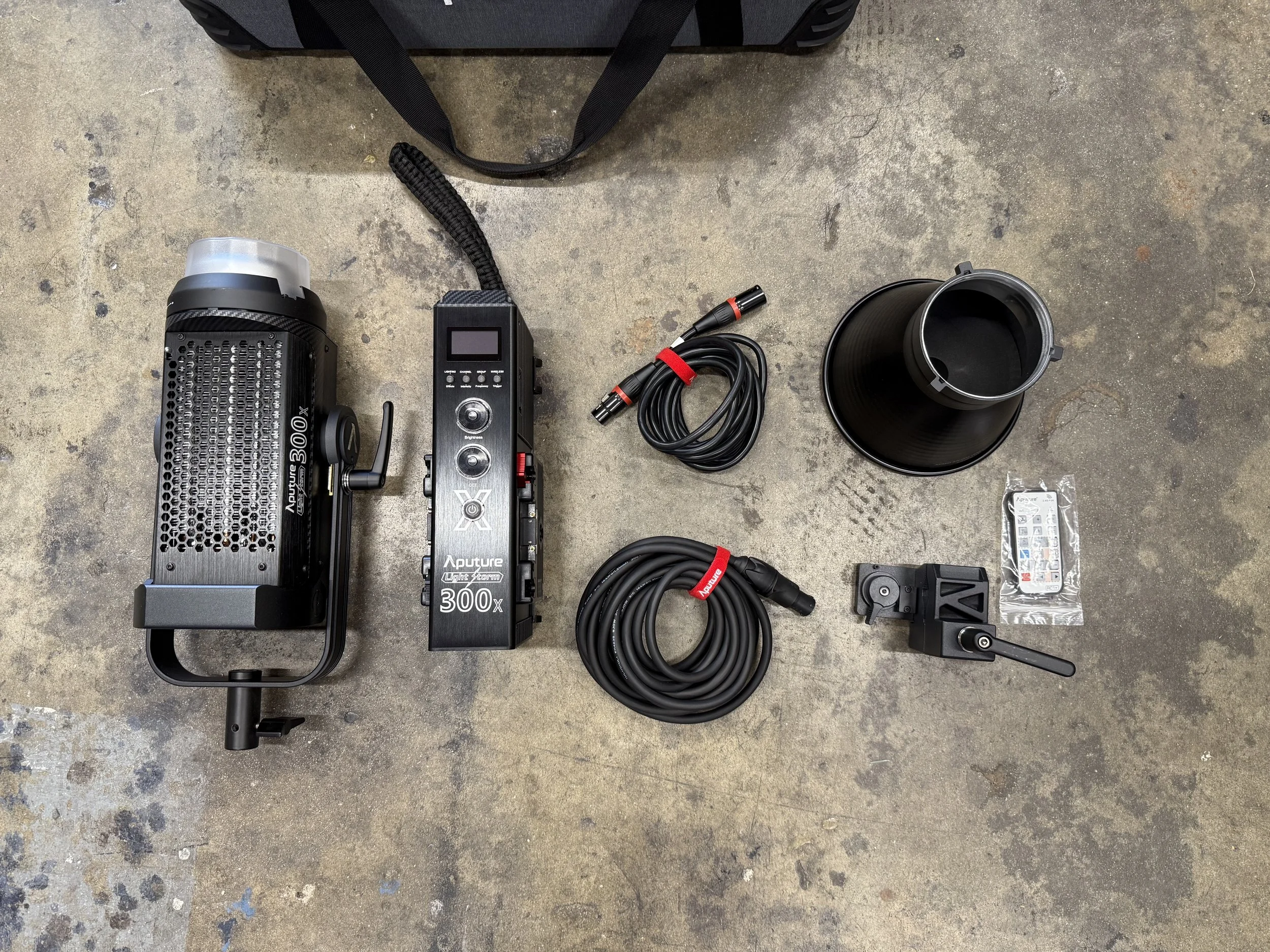 Photography of photography equipment arranged on a concrete floor. The equipment includes a studio light, a camera control unit, a power cord, a large black reflector, a remote trigger, a small clamp with a microphone, and a small pack of camera accessories, with a black bag visible at the top.
