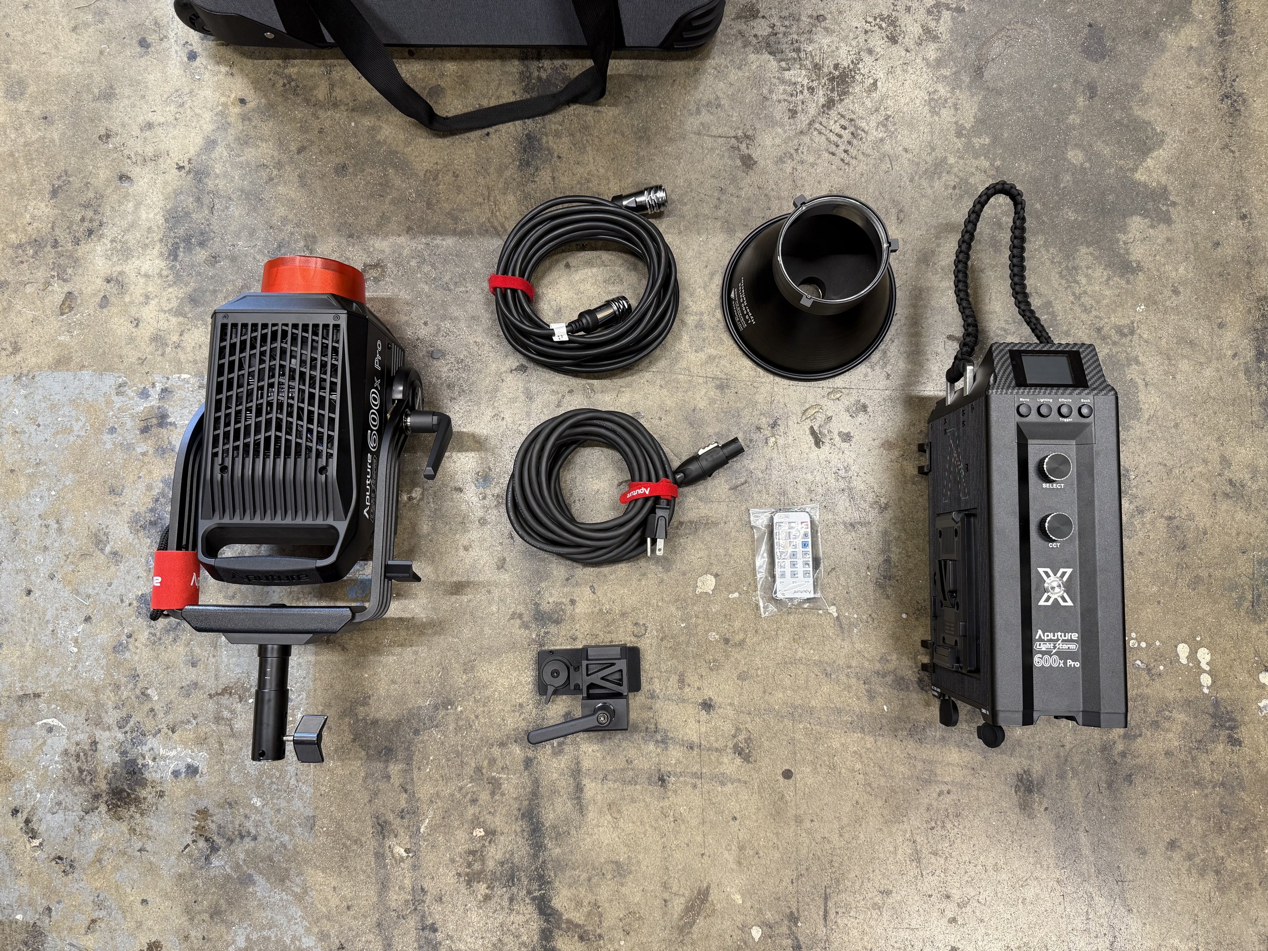 Photography of photography equipment including a large LED light, two extension cables, a black reflector, a black portable battery pack, and a small plastic bag with connectors, all laid out on a concrete floor.