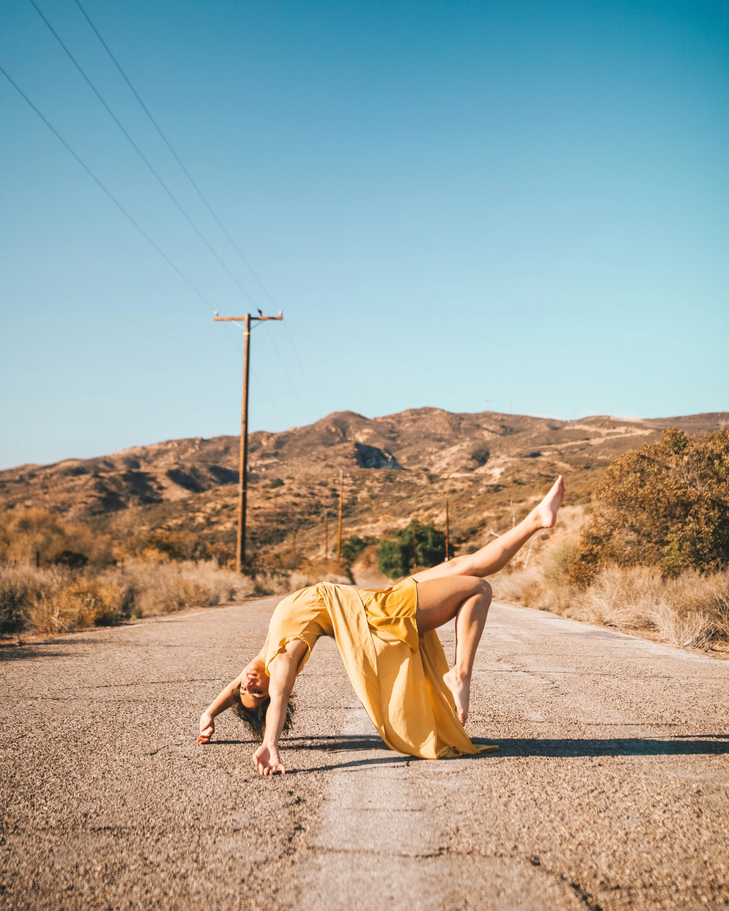 A woman in a yellow dress performing a handstand on an empty rural road with mountains in the background.