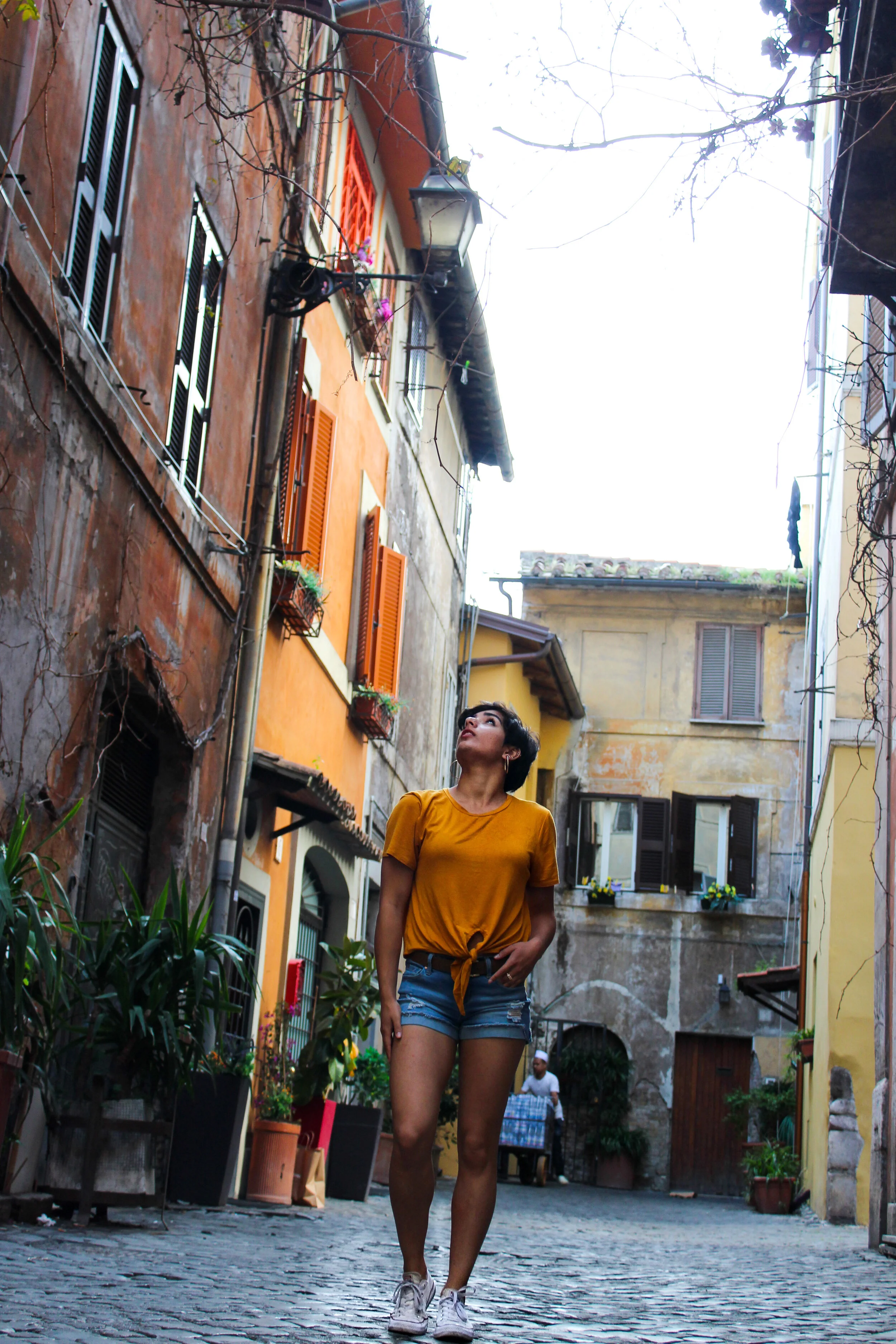 A young woman in a yellow t-shirt and denim shorts walking on a cobblestone street in a European town, looking up at the sky amid colorful old buildings with shuttered windows.