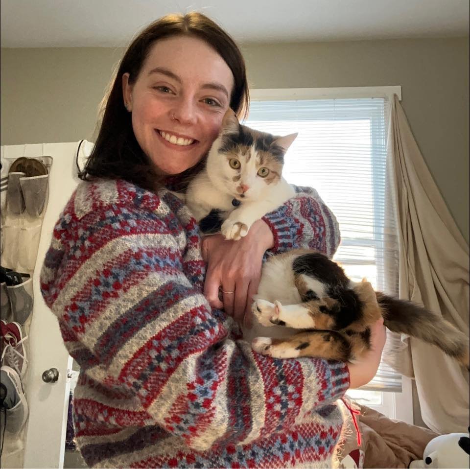 A white woman with brown hair is wearing a colorful sweater and holding a calico cat.