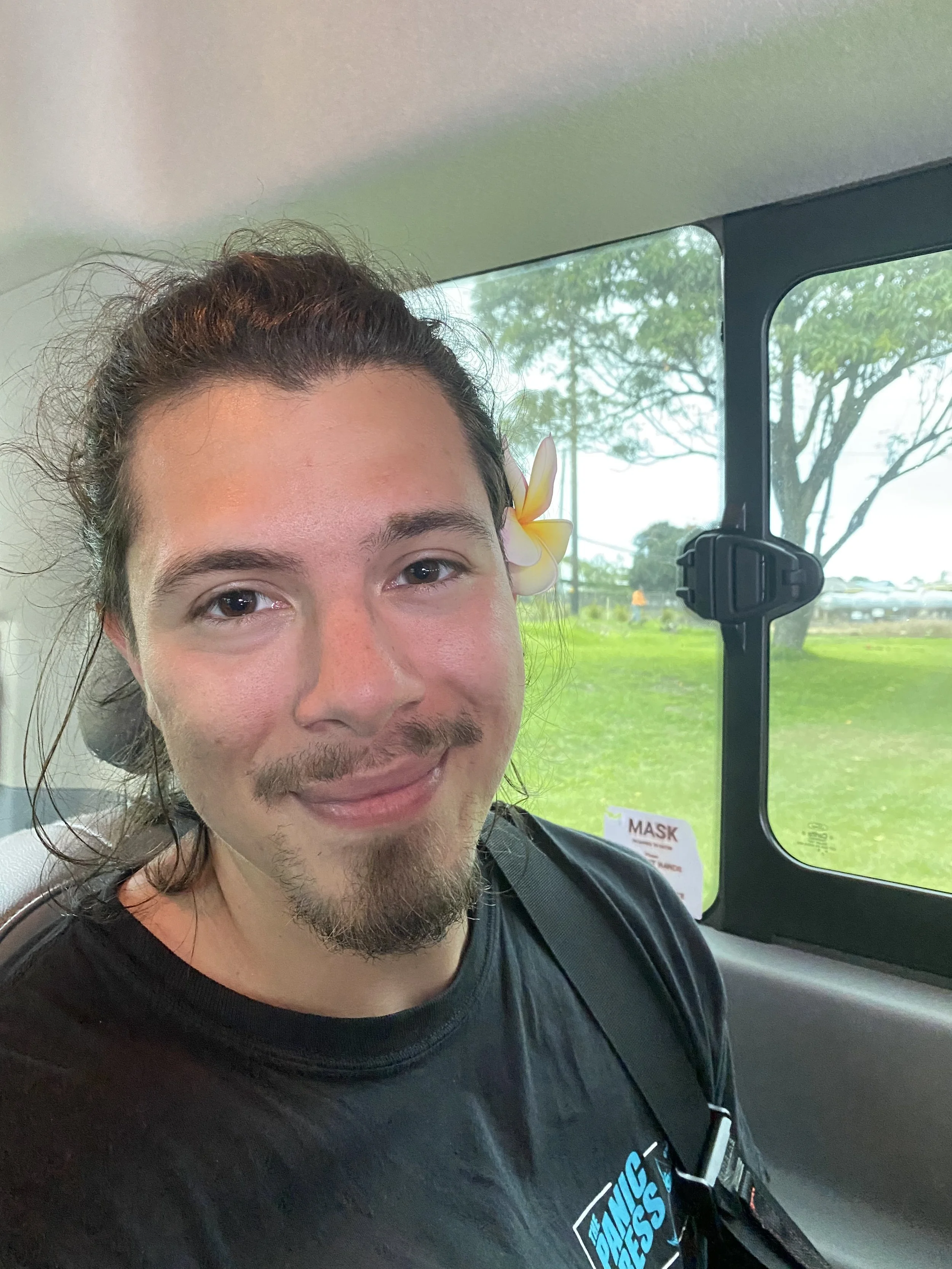 A young man with long dark hair, a mustache, and a goatee, sitting inside a vehicle, wearing a black shirt with a graphic logo, and a plumeria flower behind his right ear, smiling at the camera, with a green outdoor landscape visible through the window.