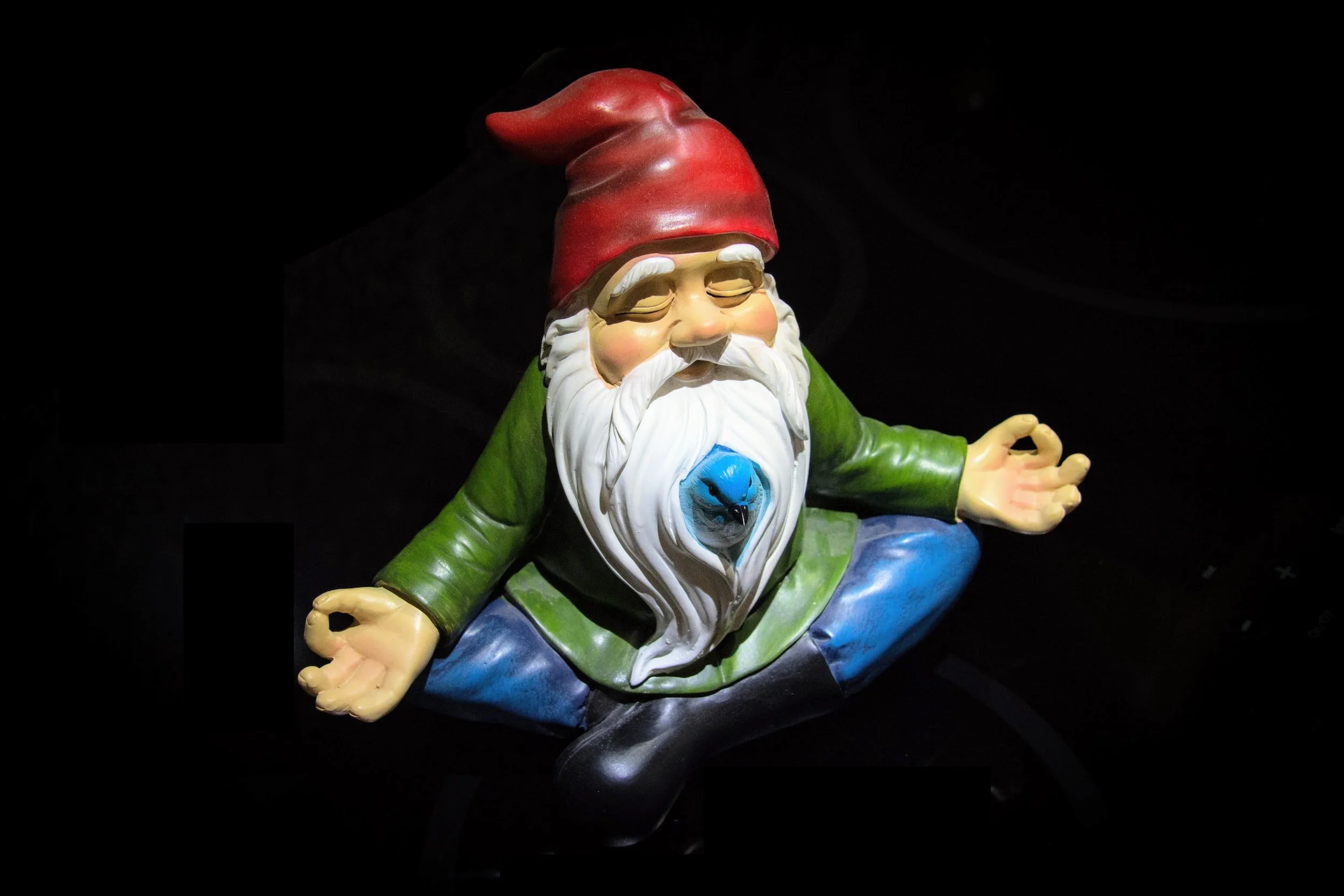 Colorful garden gnome statue in a meditative pose with closed eyes, sitting cross-legged, with hands resting on knees, against a black background.