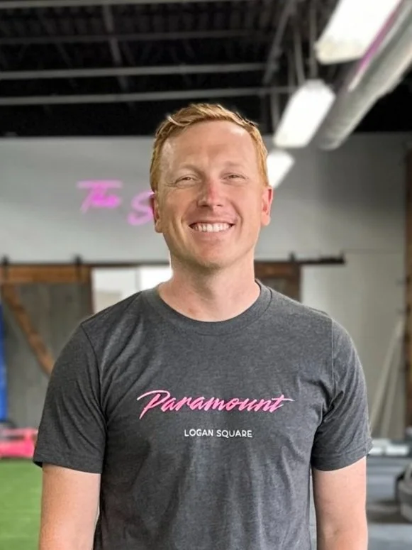 A smiling man with red hair wearing a dark gray T-shirt that has pink and white text reading 'Paramount LOGAN SQUARE'.