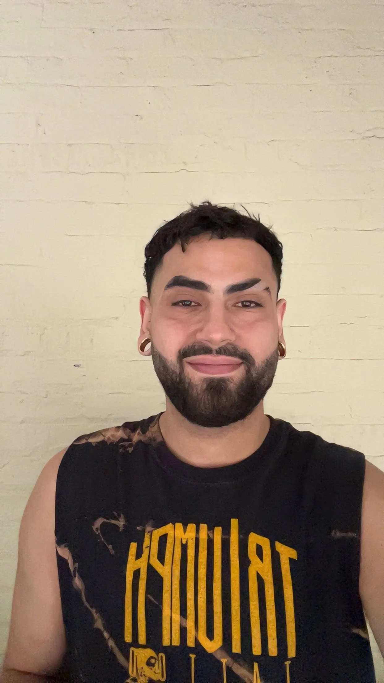 A man with dark hair, beard, and earrings smiling at the camera in front of a beige brick wall, wearing a sleeveless black shirt with yellow text.