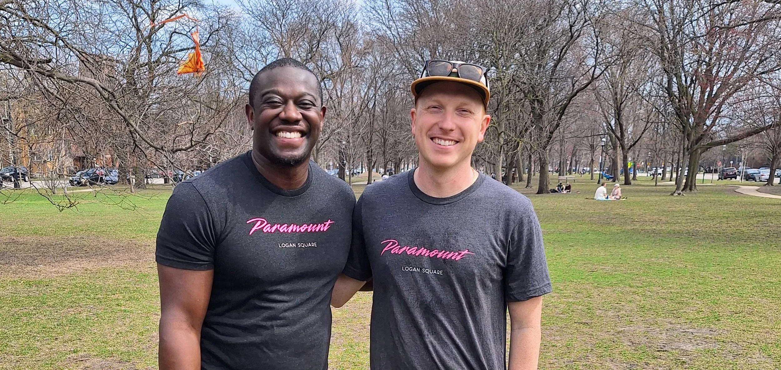 Two men standing together outdoors in a park, smiling at the camera, wearing matching gray t-shirts with pink text that reads 'Paramount Logan Square', with trees and other park visitors in the background.