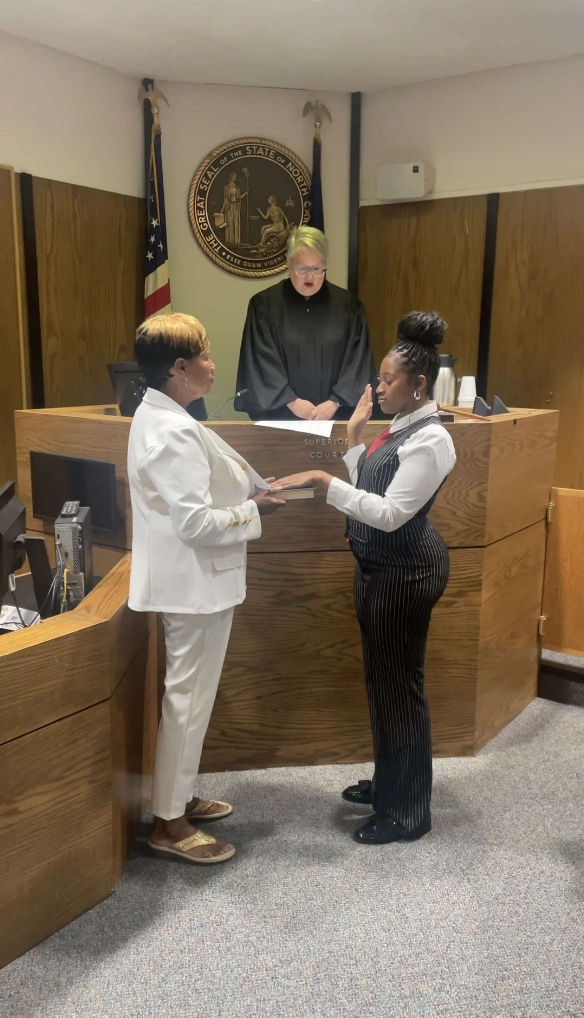 A woman in a white suit swearing in another woman in a pinstripe suit in a courtroom with a judge overseeing the ceremony.