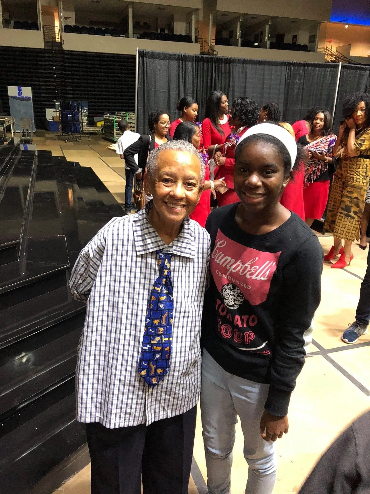 Two women smiling and posing for a photo inside a large indoor venue with a group of people in the background, some of whom are dressed in red and patterned outfits.