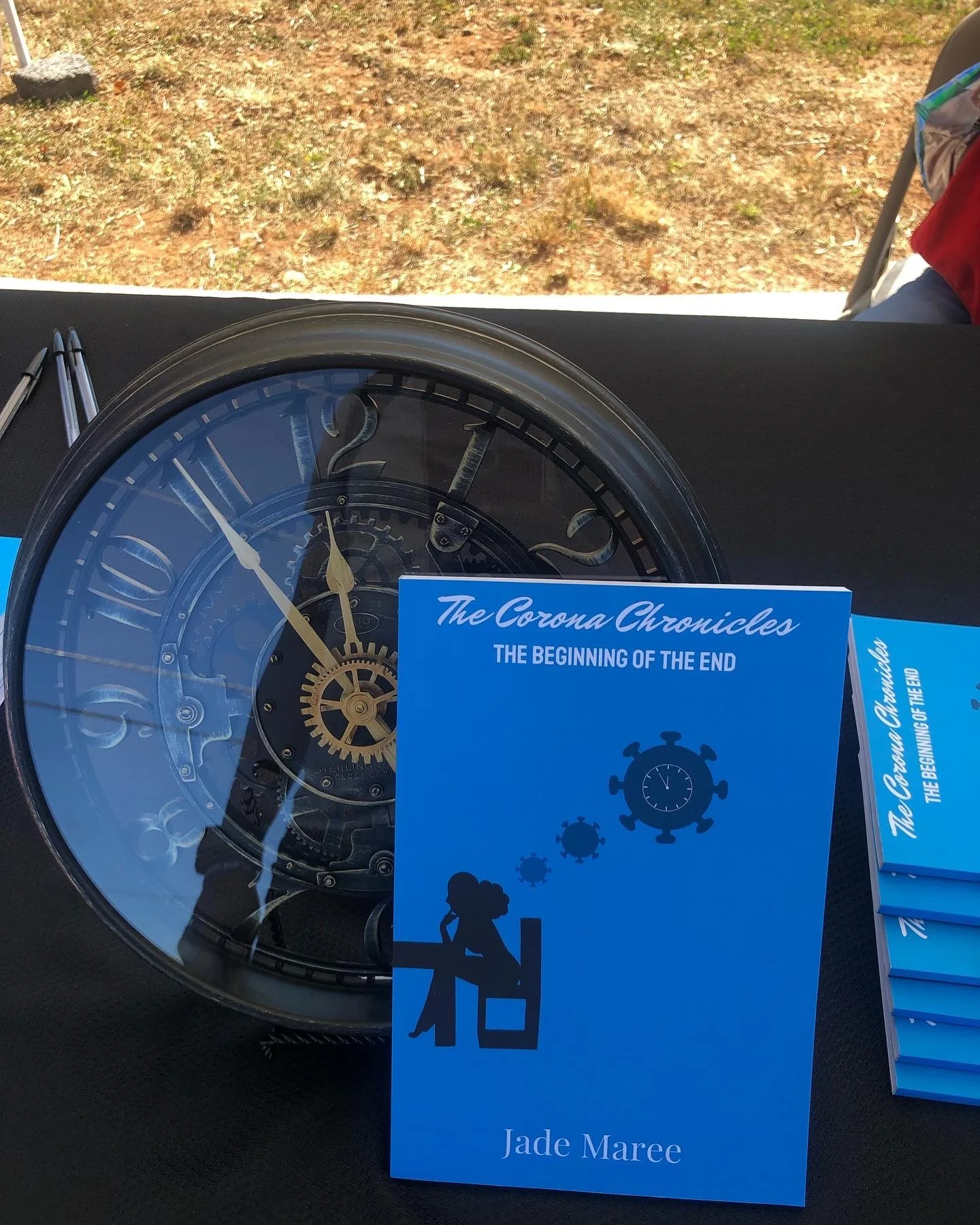 Book titled 'The Corona Chronicles: The Beginning of the End' by Jade Maree, on a table along with a decorative clock and pens, outdoors with dirt ground in the background.