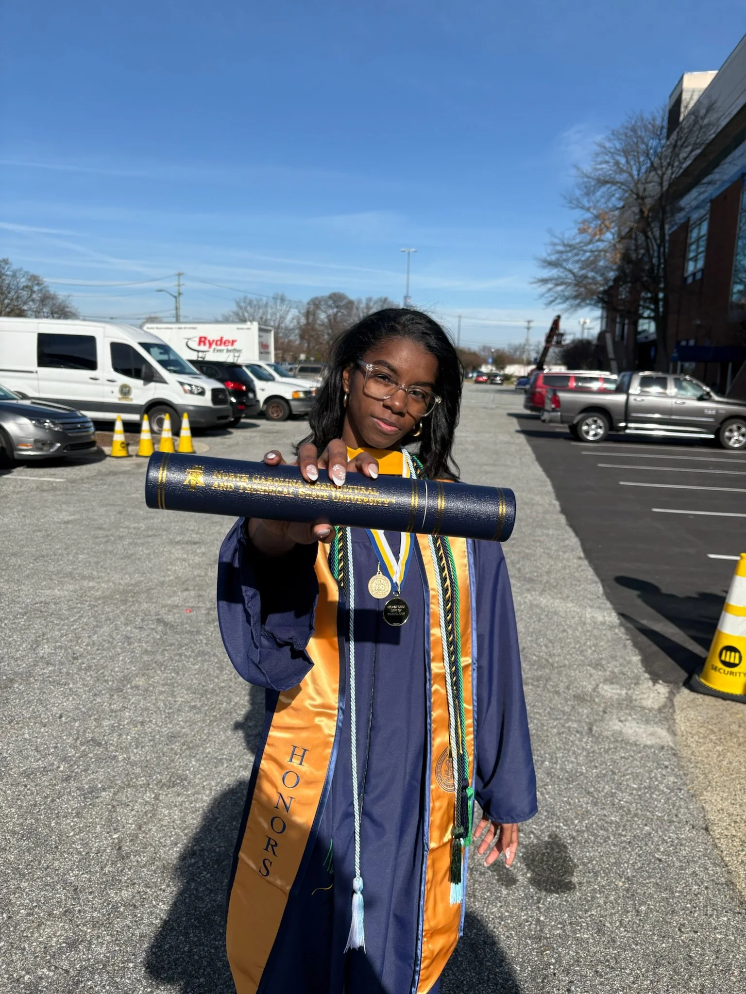 Young woman in graduation gown and honor cords holding diploma in a parking lot.