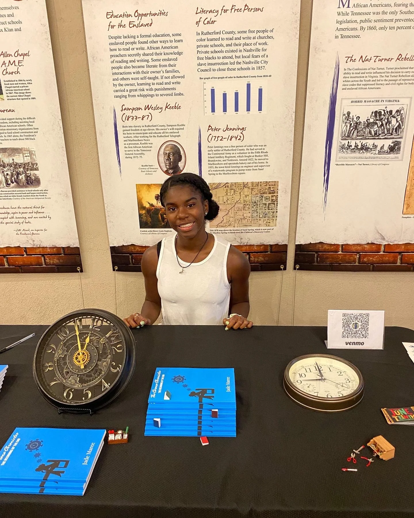 Young girl standing at a table with clocks, books, and small toys, smiling at camera, with informational posters about historical African American figures and history in the background.