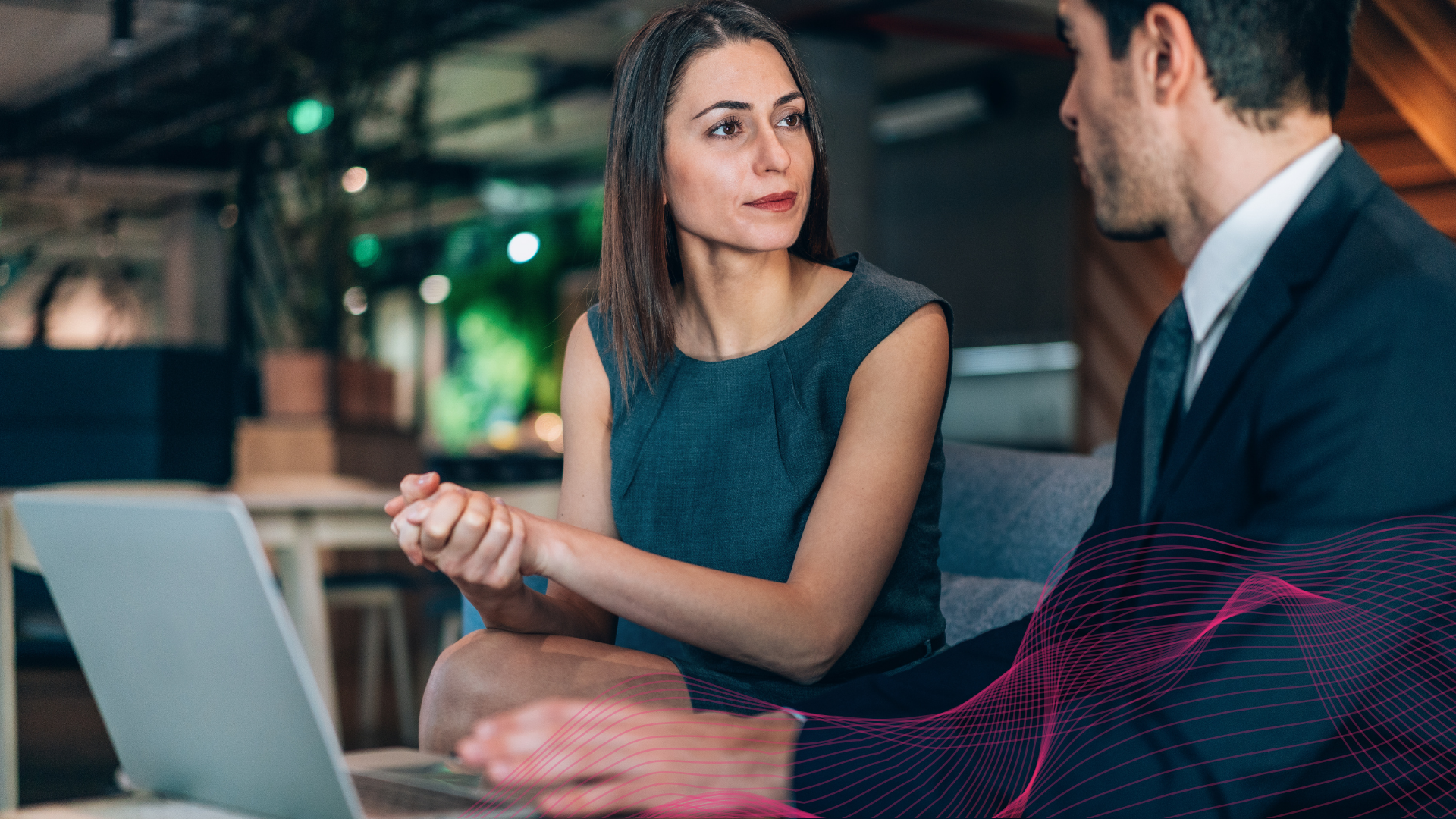 A woman in a sleeveless gray dress engaged in a conversation with a man in a suit, sitting in a modern, dimly lit workspace or office, with a laptop open in front of her.
