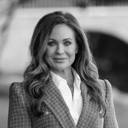 A woman with long wavy hair wearing a checkered blazer and white shirt, standing outdoors with blurred background.