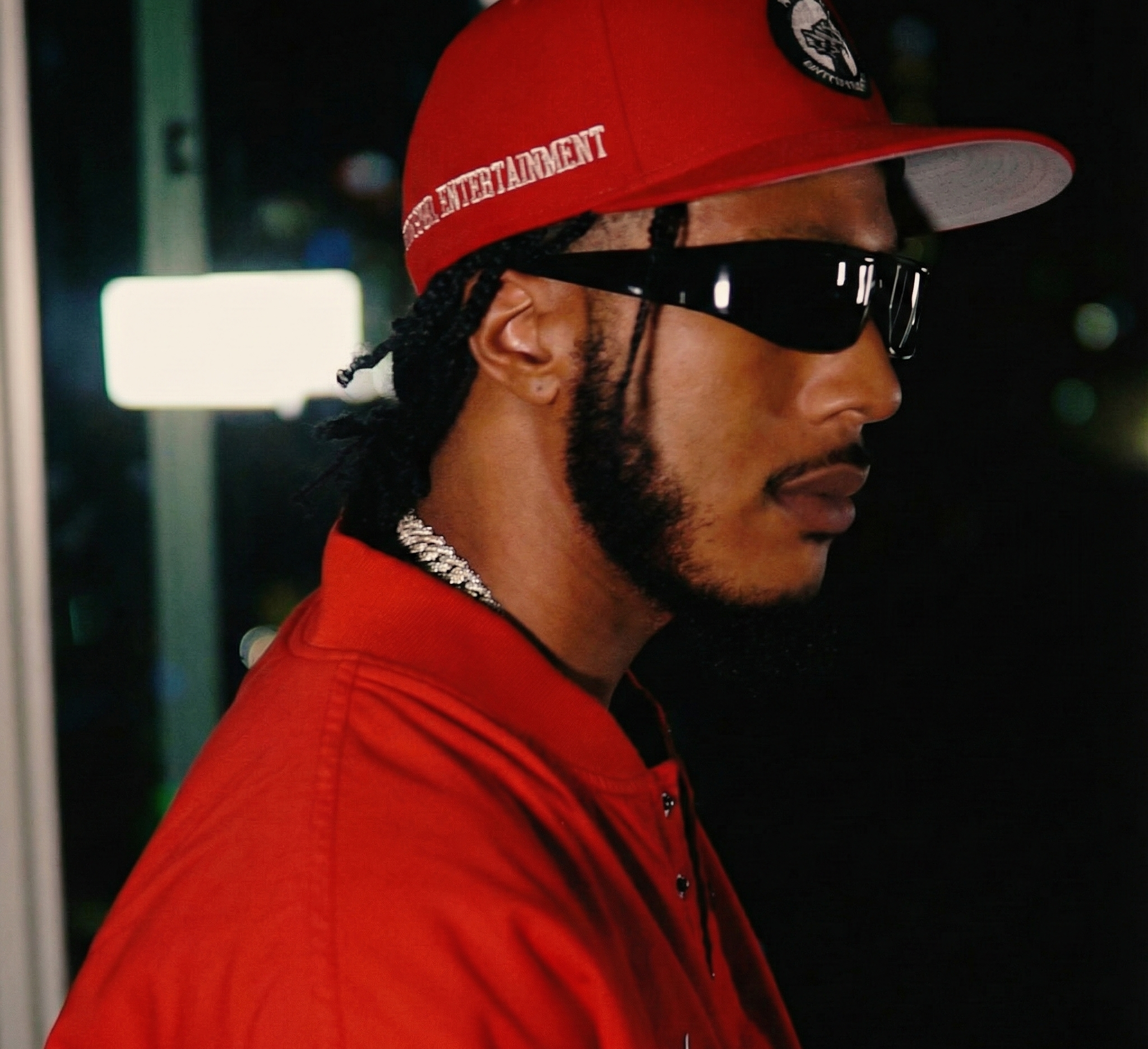 Side profile of Markel X.L. with dreadlocks wearing a red cap with 'ENTERTAINMENT' written on it, black sunglasses, and a red shirt, standing at night with blurred city lights in the background.