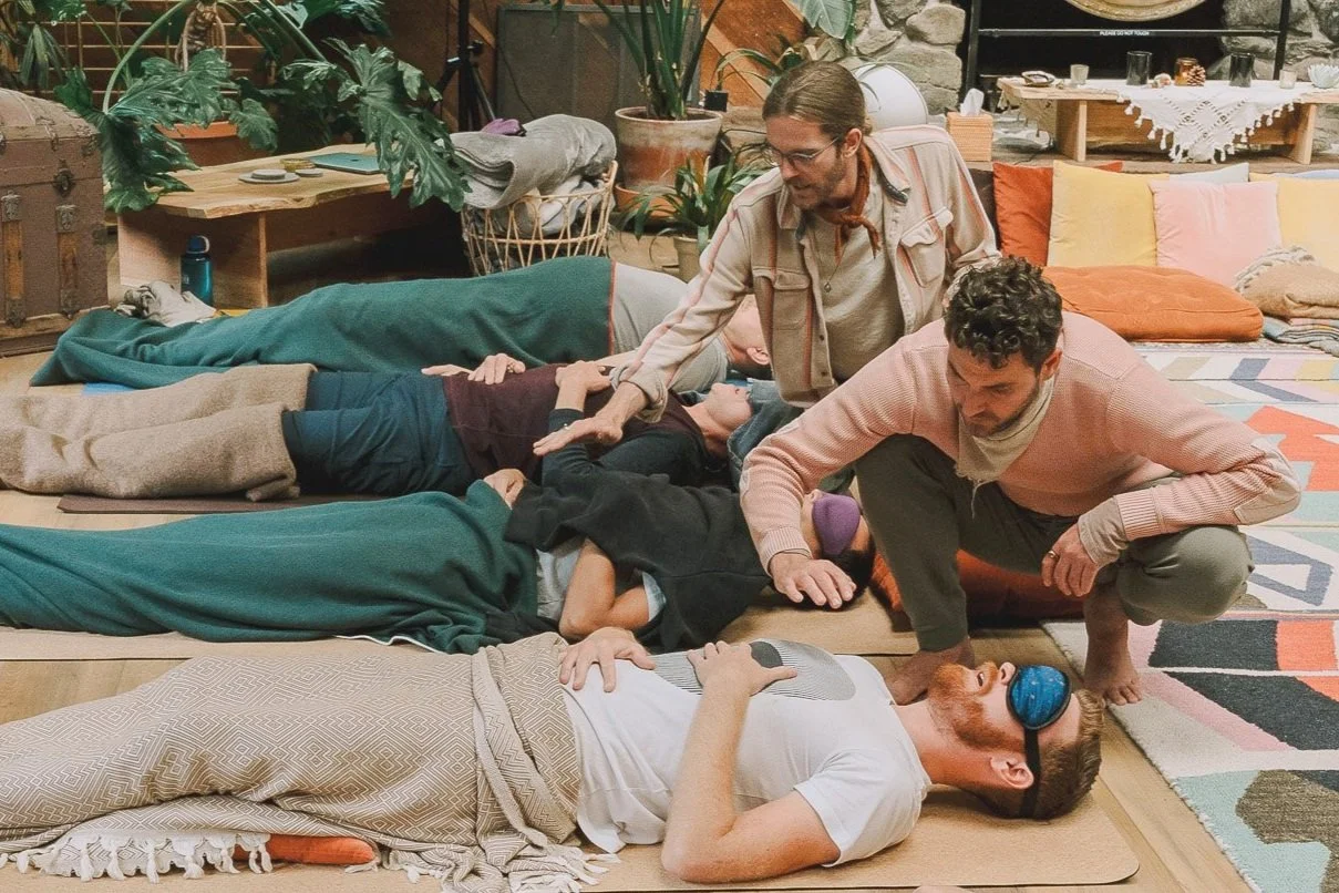 A group of people lying on the floor and one person hypnotized with a sleep mask, while two others appear to be performing a hypnotism or rescue process in a cozy, decorated room with plants and colorful cushions.