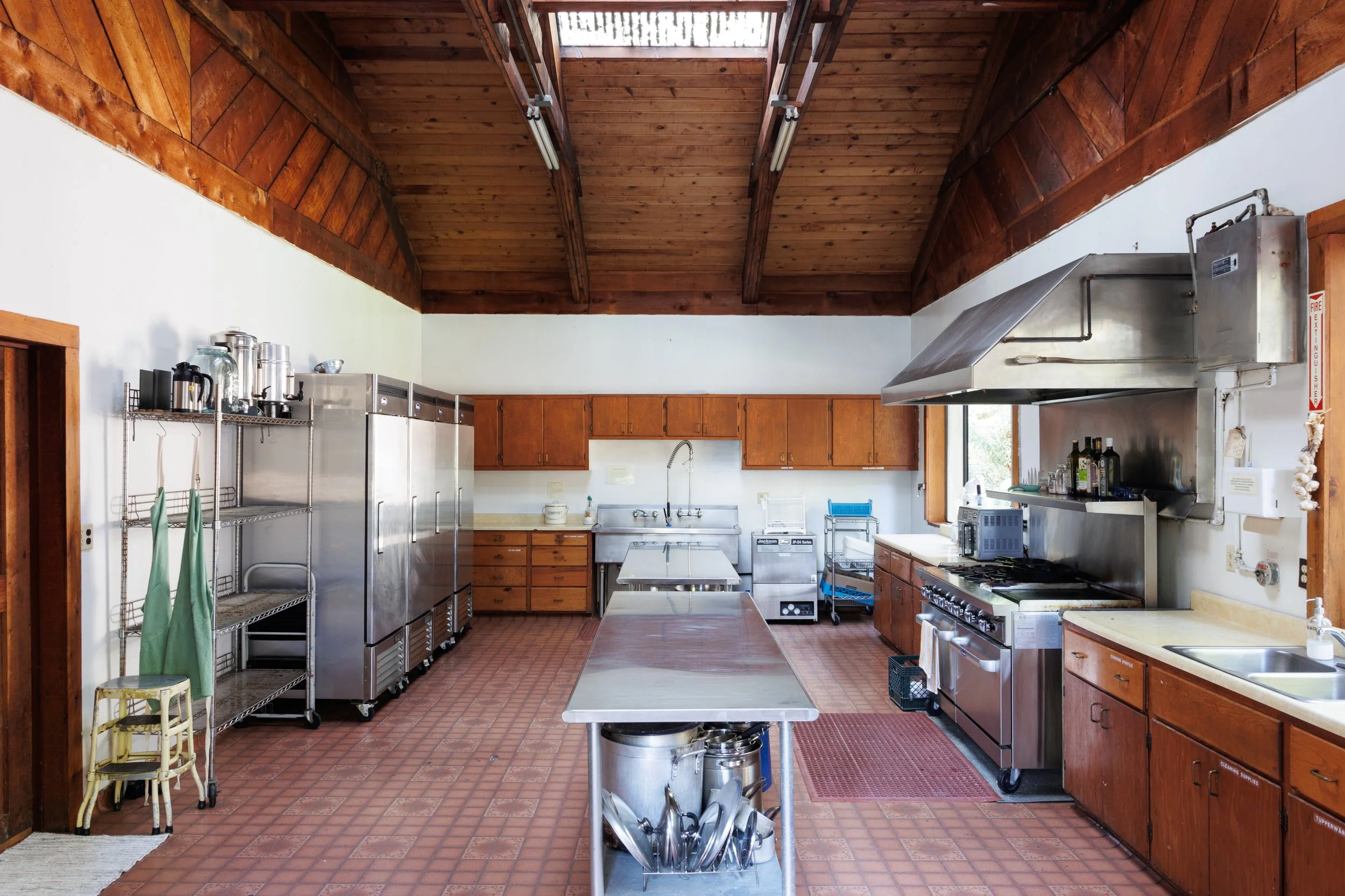 Commercial kitchen with wooden ceiling, stainless steel appliances, and a central metal prep table.