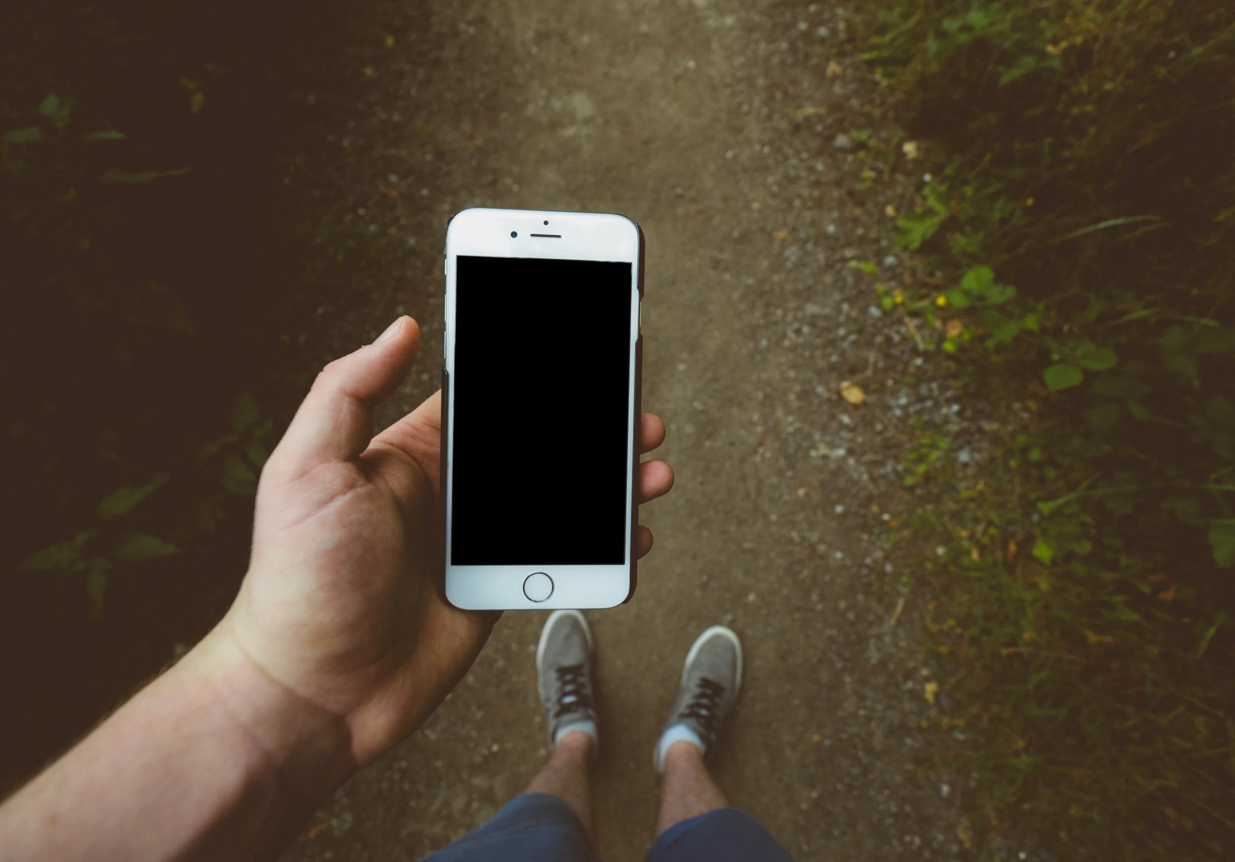 A person holding a smartphone with a black screen, standing on a dirt trail surrounded by green foliage, with their legs and sneakers visible.