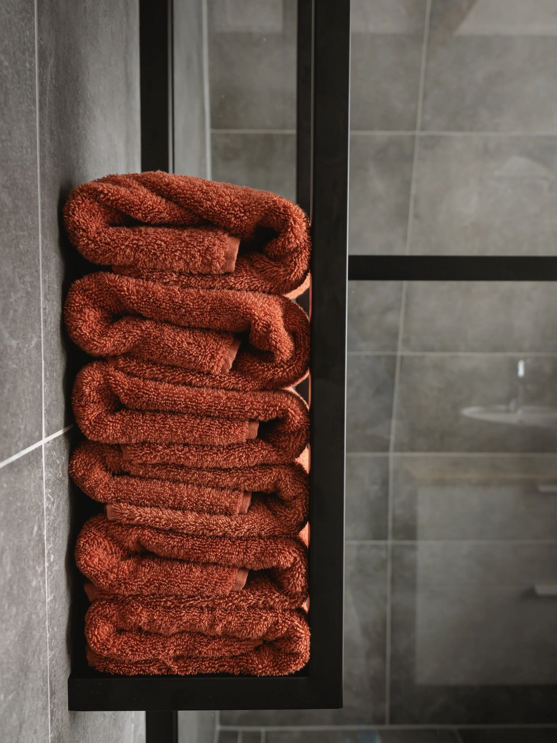 Stack of six folded, brownish-orange towels on a black towel rack in a bathroom.
