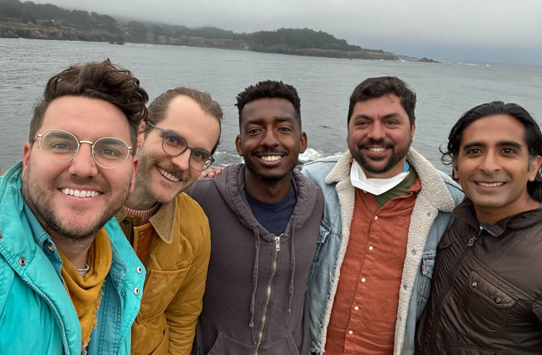 Five men standing close together on a beach, smiling at the camera, with the ocean and a cloudy sky in the background.