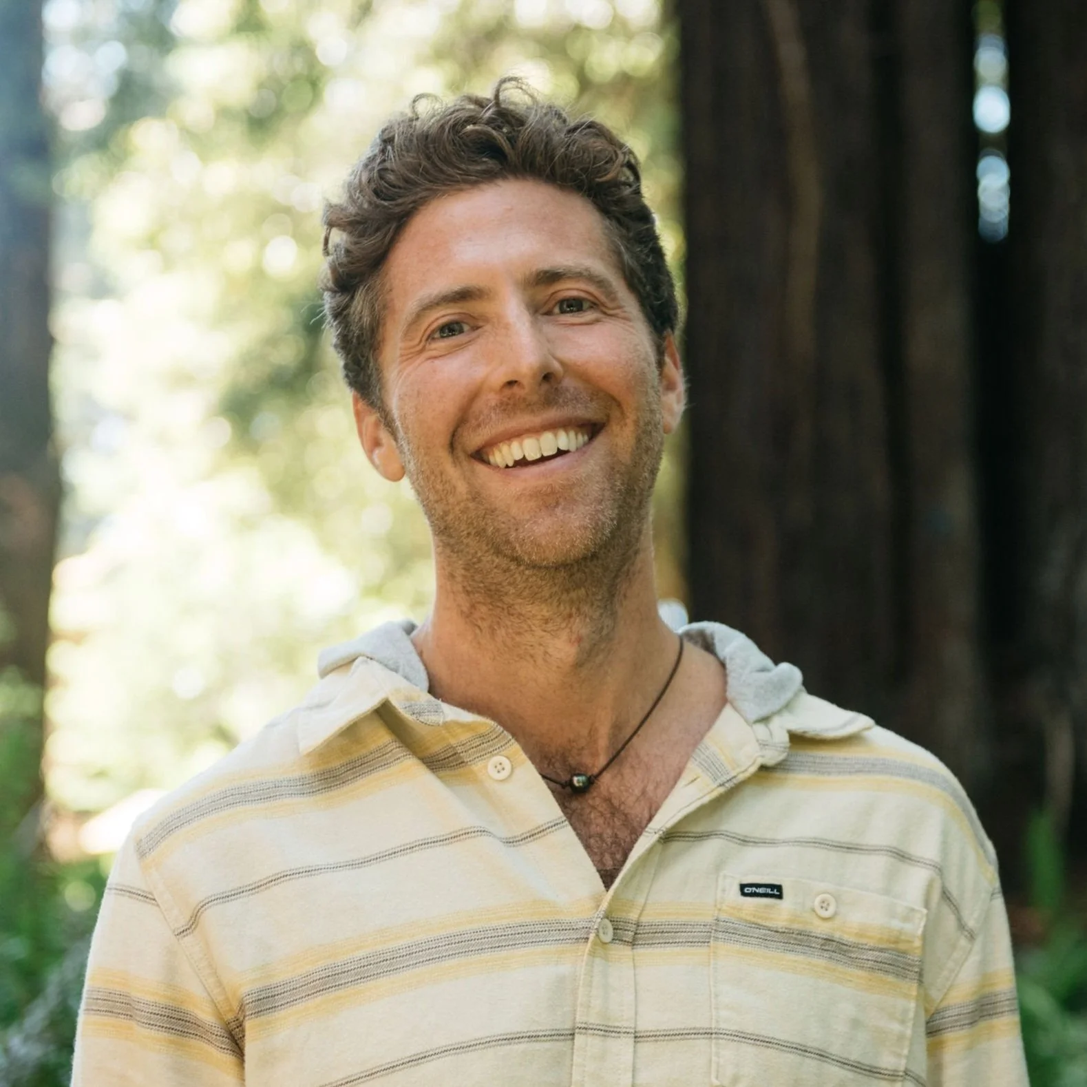 A smiling man with curly hair, wearing a light yellow striped shirt and a black necklace, outdoors with green trees in the background.