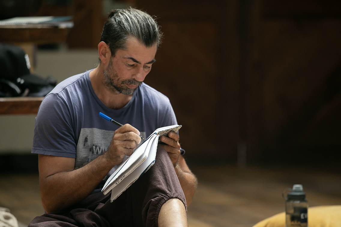 A man with dark hair and a beard, wearing a blue t-shirt, sitting cross-legged and writing in a notebook with a blue pen in a wooden room, with a water bottle nearby.